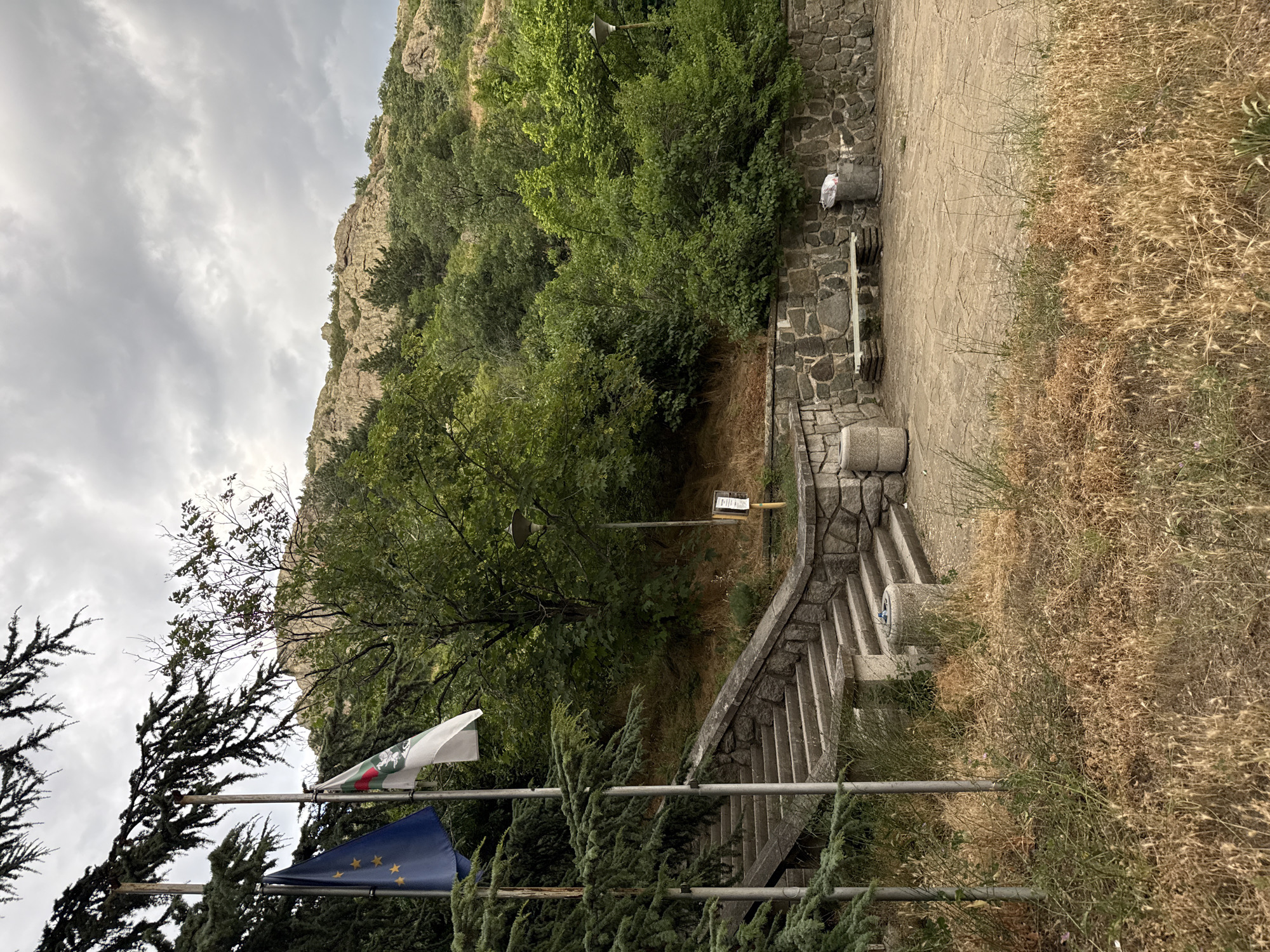 The image depicts an outdoor scene in Sliven, Bulgaria, with a focus on a set of stone stairs leading upwards. Two flags are visible on flagpoles to the left of the stairs: a blue European Union flag and a white flag with a green and red stripe and a crest. The stairs are flanked by dense green foliage, including coniferous trees on the left. Behind the stairs, the landscape rises, covered in trees and rocky outcrops, characteristic of the Sliven area. In the foreground, dry grasses and weeds cover the ground. To the right of the stairs, a stone wall supports a paved area with a bench, two cylindrical trash cans, and a lamppost. The sky is overcast with grey clouds. No people are present in the scene.