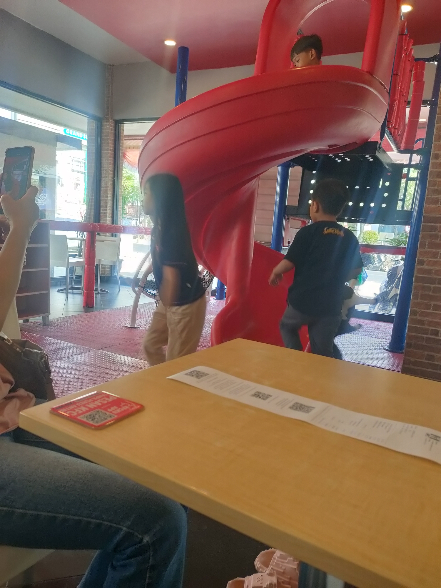 The image depicts an indoor play area, likely at a fast-food restaurant, as evidenced by the presence of a red plastic slide structure and a table with receipts and a QR code ordering sticker. Two young boys are visible on the play structure, one near the top of the slide and another on the platform below. A person is seated at the table, with their legs in jeans and a portion of their arm visible, holding a smartphone. The environment is brightly lit, with red and blue accents on the play equipment and a brick wall in the background. The flooring is a red, textured mat. The overall scene suggests a family outing or a visit to a restaurant with a children's play area in Subang, Indonesia.