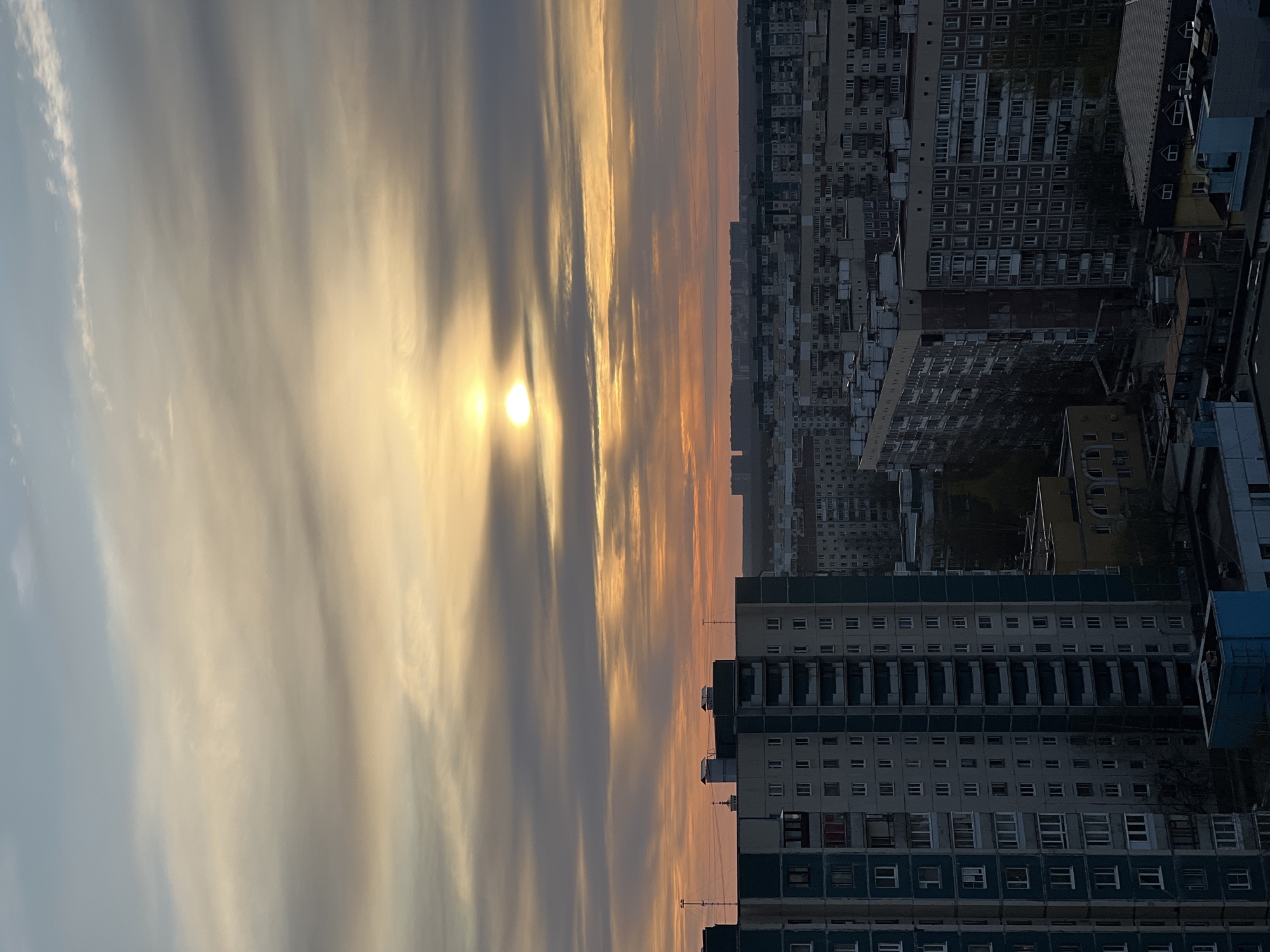 The image captures a sunset over Saint Petersburg, Russia. The sun is partially obscured by clouds, creating streaks of light in the sky. Below the sky, a dense urban landscape unfolds, featuring multiple high-rise apartment buildings. The buildings are typical Soviet-era constructions, marked by repetitive window patterns. The lighting suggests that the photo was taken during the late afternoon or early evening.