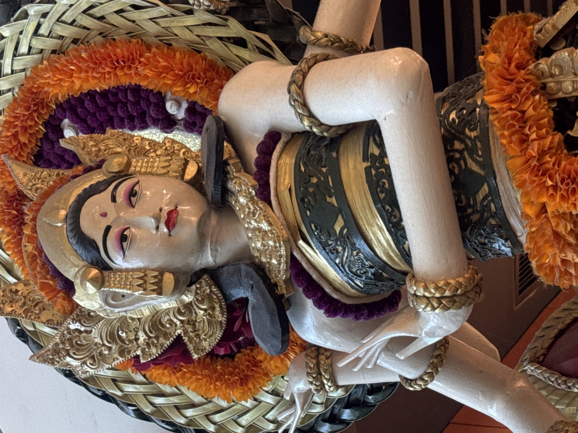 A close-up, slightly angled shot depicts a Balinese sculpture, likely photographed in Kuta, Indonesia. The sculpture is adorned with vibrant garlands of orange and purple flowers, set against a woven basket-like background. The figure has fair skin, defined facial features, and is elaborately dressed with gold jewelry and decorative accents. The sculpture's hand is visible, adorned with a braided gold bracelet. A black and gold patterned element wraps around the body. The overall scene creates a rich, cultural aesthetic.