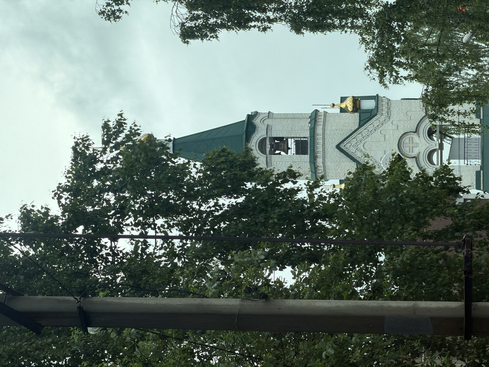 The image displays a partial, eye-level view of the Church of the Blessed Matrona of Moscow in Votkinsk, Russia, under an overcast sky. The primary subject is the church's white brick bell tower, topped with a steep, conical green roof culminating in a small golden onion dome. A large bell is visible within one of the bell tower's arched openings. Below the bell tower, additional architectural elements include white brick facades, arched windows, and a black metal railing on a lower section of the building. Several smaller golden domes are situated on other visible sections of the church roof.
In the left foreground, a substantial grey concrete utility pole stands vertically, partially obscuring the church. Wires are attached to the pole, and a translucent, light-colored object, possibly an insulator, is visible on its left side. Dense green foliage from multiple trees frames the church on both the left and right sides, partially obscuring the view of the structure and the sky. Another dark, slender vertical element, likely a thinner pole or tree trunk, is visible within the foliage to the right of the concrete pole. The sky overhead is a uniform light grey, indicating heavy cloud cover. No individuals or animals are present in the scene, and no explicit actions or interactions are depicted. The overall environment suggests an urban or suburban setting.