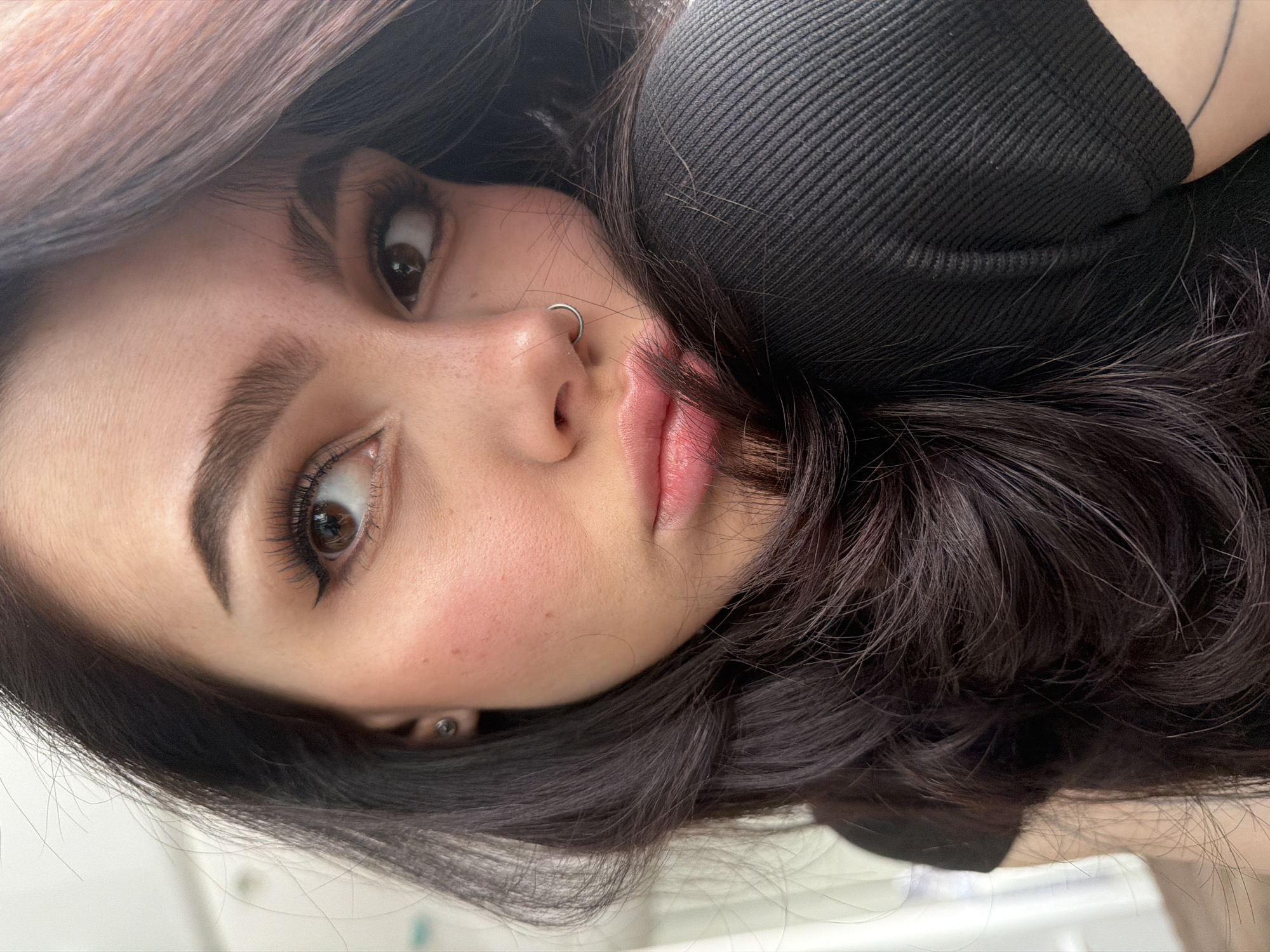 A close-up portrait features a young person, likely female, with dark, wavy hair, looking directly into the camera while reclining or lying down. The image is rotated, orienting the subject's head to the right. She has large, dark brown eyes accentuated with eyeliner and mascara, and thick, well-defined dark eyebrows. Her nose is small, featuring a silver hoop piercing on the left nostril. Her lips are full and tinted pink. Her fair skin shows subtle rosy blush on the cheeks and faint freckles. Dark brown hair, possibly with subtle lighter strands, frames her face and flows around her. She wears a black ribbed top, visible on the upper right. A small stud earring is visible on her right earlobe, partially obscured by her hair. The background is softly blurred and light-colored, indicating an indoor setting. The lighting is soft and even, highlighting her features. No text is visible.