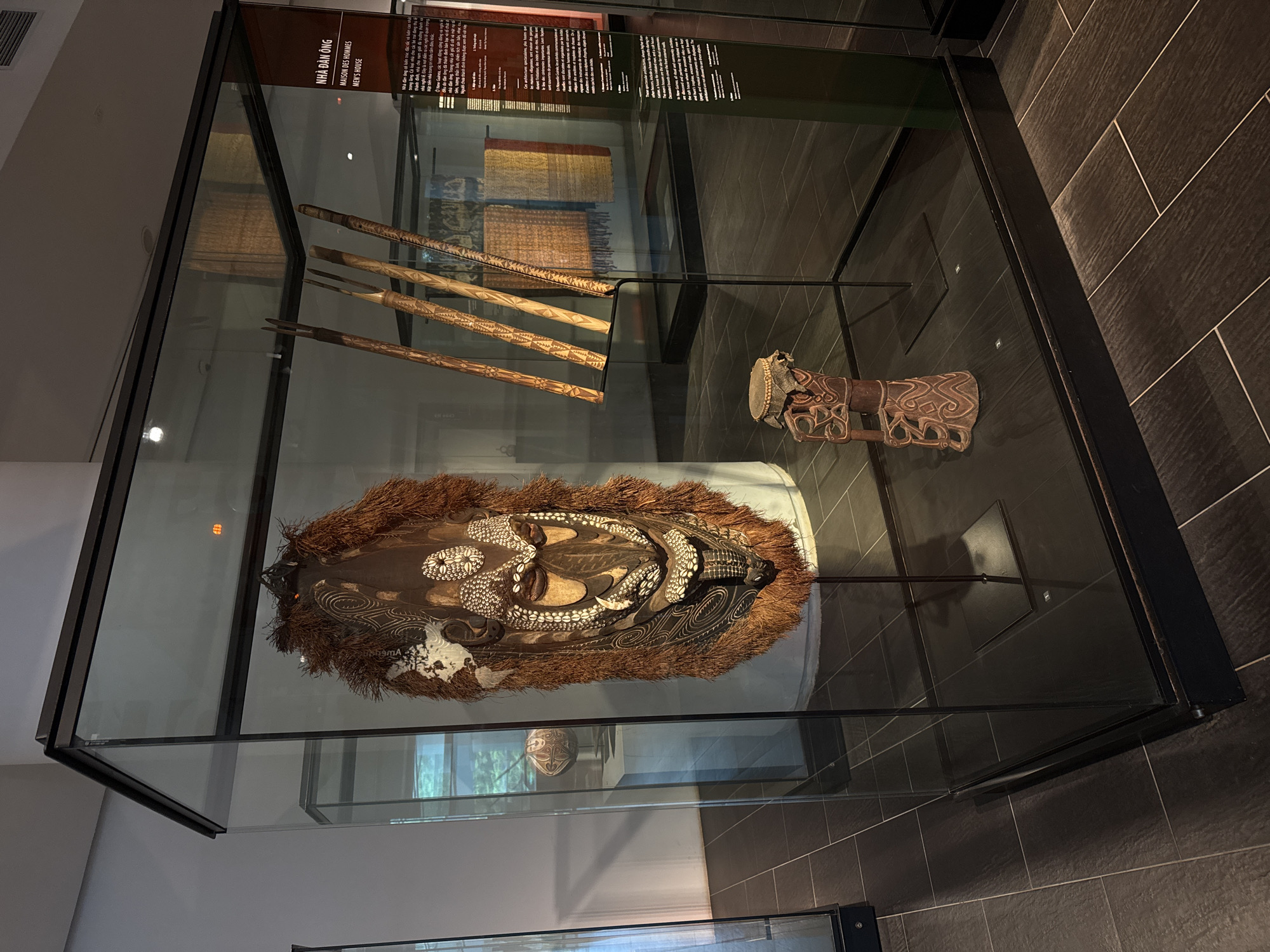 This media file captures an exhibit in a museum, likely located in Hà Nội, Vietnam, as suggested by the Vietnamese text visible. The primary subject is a display case containing cultural artifacts. Inside the case, a large, ornate mask with a fur trim is prominently featured. The mask is intricately decorated with shells and carved patterns. To the left of the mask, several long, carved wooden objects are displayed vertically, leaning against the back of the case. On the right, a small drum and a carved wooden figure are also exhibited. Behind these objects, more fabric artifacts are visible, appearing to be textiles with woven patterns in red, yellow, and brown hues.
The display case is modern, made of glass and black metal, and is illuminated, suggesting an indoor museum setting. The background includes dark tiled walls, characteristic of many contemporary museum designs. The text on the upper left of the display case, partially visible, is in Vietnamese and French, indicating the exhibit's cultural context. The visible Vietnamese text translates to "Men's House," suggesting the artifacts are related to male cultural practices or spaces. The overall atmosphere is one of quiet observation and cultural preservation. The lighting is artificial and focused on the display, creating a contemplative mood.