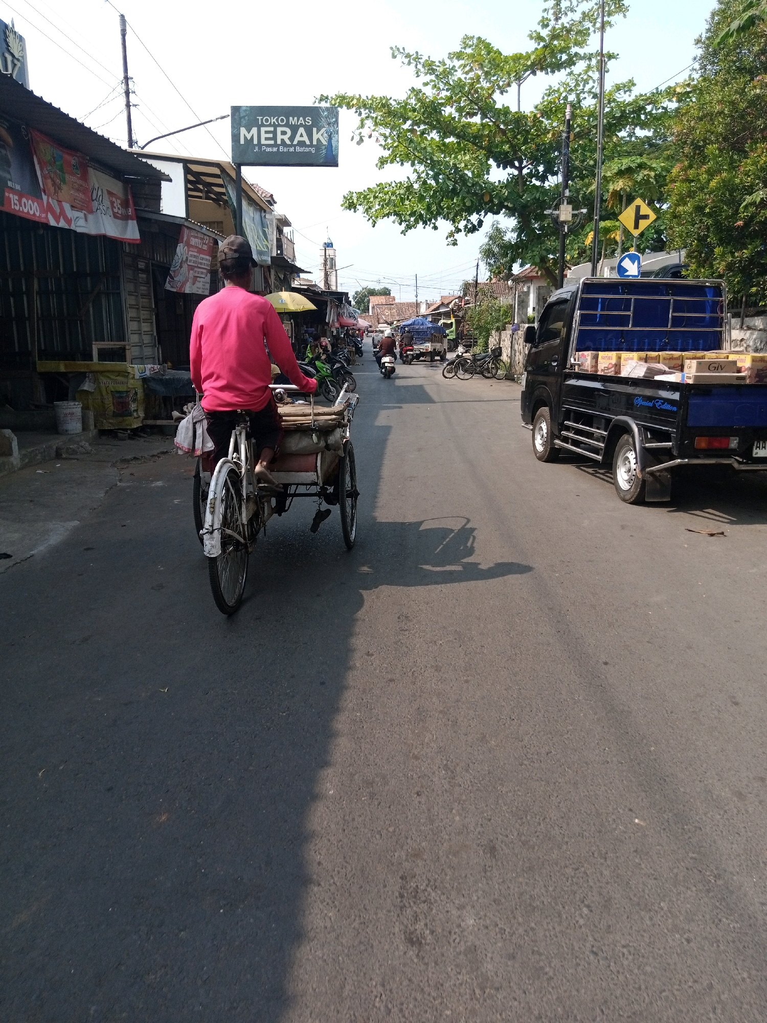 In Proyonanggan Tengah, Indonesia, a man wearing a bright pink long-sleeved shirt rides a tricycle cart on a paved road. The tricycle has a white frame and two wheels at the back, one of which is larger and more prominent. The man is seated and appears to be pedaling. The cart behind him contains some items covered by fabric. The road is lined with businesses on the left, displaying banners and awnings. A sign for "TOKO MAS MERAK" (Merak Gold Shop) is visible, along with other advertisements. On the right side of the road, a blue pickup truck is parked, its bed loaded with boxes. The environment suggests a commercial area with a mix of old and newer buildings, trees, and overhead wires. The sky is clear and bright, indicating daytime. Other vehicles, including motorcycles, are also present on the road in the distance.