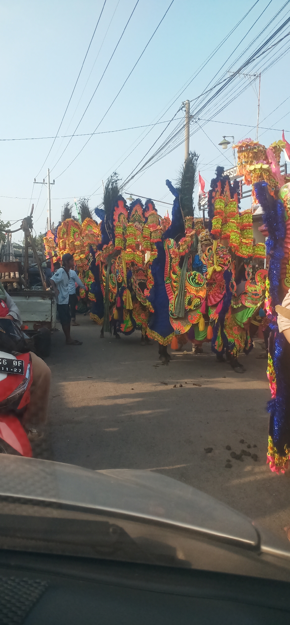 A street scene in Kalisalam, Indonesia, is captured from the perspective of a vehicle, showing a procession of people in elaborate, colorful costumes. The costumes appear to be inspired by traditional Indonesian cultural performances, possibly depicting mythical creatures or characters, adorned with bright fabrics, tassels, and ornate headdresses with feather-like accents. The costumes are predominantly blue, red, yellow, and green, with intricate golden detailing.
Several individuals are visible. In the foreground on the left, a person is seated on a red and white motorcycle. To the left of the procession, a man in a light blue short-sleeved shirt and dark shorts stands facing the costumed figures. His hands are in his pockets. Behind him, the back of a white truck is visible, loaded with what appear to be bamboo poles.
The street is paved and shows signs of being a public thoroughfare. In the background, utility poles with a dense network of wires crisscross the sky, indicating an urban or semi-urban environment. The sky is clear and pale blue, suggesting daylight. Sunlight casts shadows on the road, indicating the time of day is likely late afternoon or early morning. Scattered on the ground are small dark clumps, possibly animal droppings. The overall impression is a vibrant cultural event taking place on a typical street.