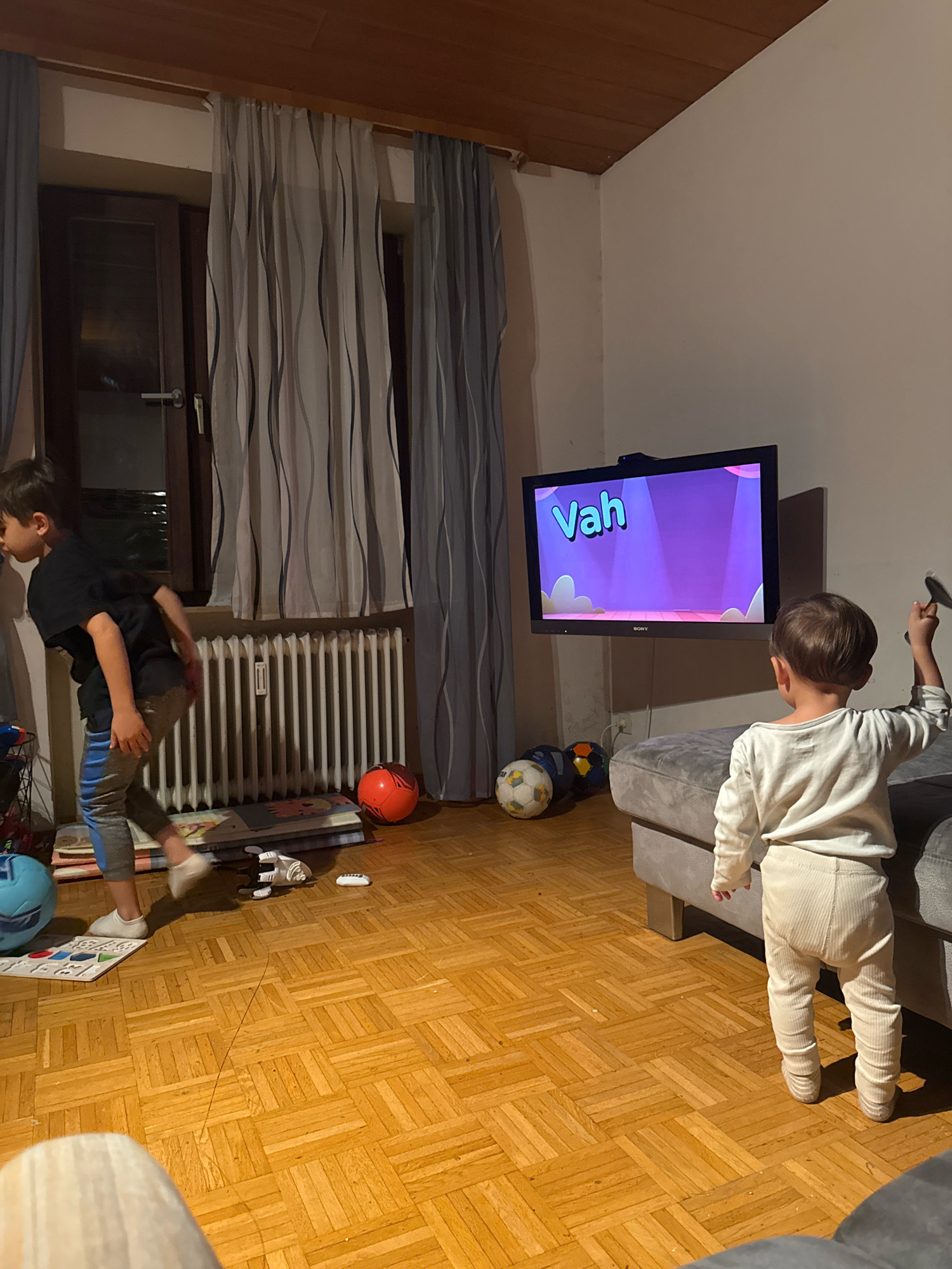 In a living room in Überherrn, Germany, two young children are engaged in activities. On the left, a boy wearing a black t-shirt and grey and blue striped pants stands facing a window, with his back to the camera, his left hand near his hip. In front of him, on the wooden floor, are several items including a light blue ball, a white and black toy, and some books or mats. A radiator is visible behind him. On the right side of the room, a second, younger child, clad in a white long-sleeved shirt and beige ribbed pants, stands facing a television screen. The television, mounted on the wall, displays a purple screen with stylized white clouds and the word "Vah" in large yellow letters. Two soccer balls, one white and black, and one dark blue and yellow, are on the floor near the child. A grey sofa is partially visible behind the second child. The room is lit by artificial light.
