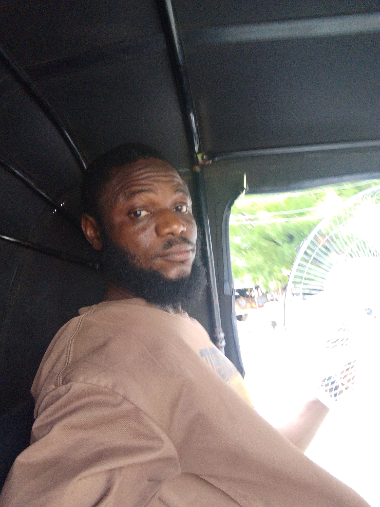 A medium shot captures a man with dark skin and a black beard inside a vehicle, potentially a tuk-tuk, given the metal frame visible above him. He is wearing a light brown shirt and has a tattoo on his left arm. The man is looking directly at the camera. Through the window on his right, a blurred street scene is visible, including a white fan, vegetation, and potentially some buildings or shops, suggesting the location is in an urban area. Given the prompt, the location is likely Adankolo, Nigeria.