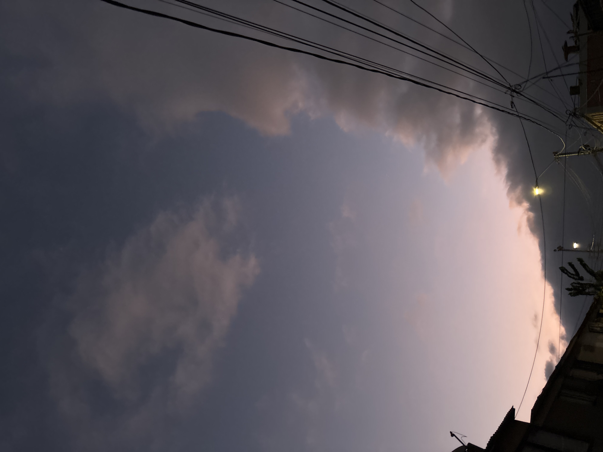 An upward-angled shot shows a cloudy sky at dusk, punctuated by power lines and the corner of a building in Arica, Chile. Streetlights are visible along the right edge of the image. The sky transitions from a dark grey to a lighter pinkish hue.