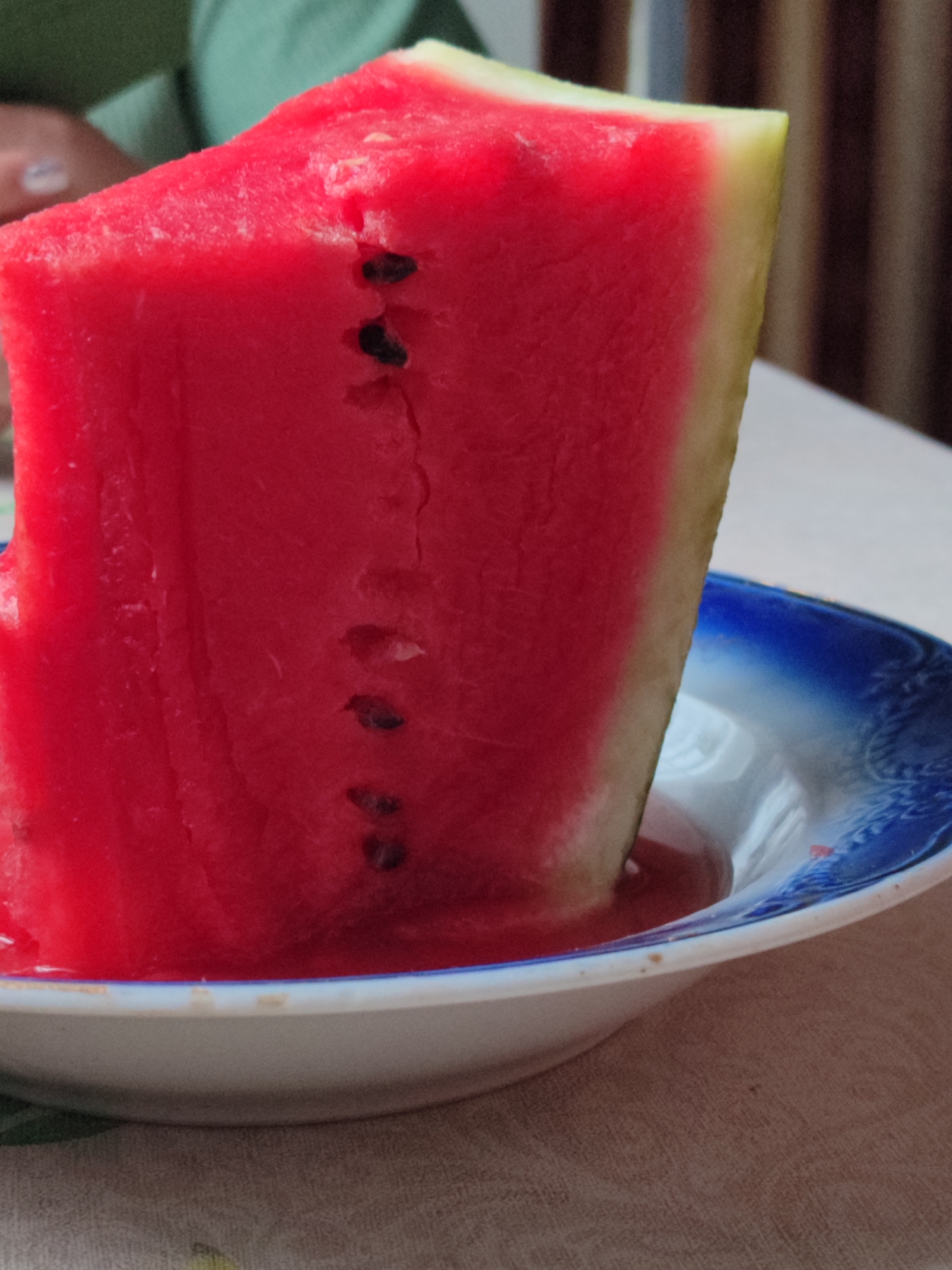 A slice of watermelon sits in a white and blue patterned bowl on a tan tablecloth. The watermelon is bright red with a yellow rind and several black seeds. A person wearing a green shirt is partially visible in the upper left corner of the image, with their hand resting on the watermelon. The background is blurred and appears to be an indoor setting with vertical blinds.