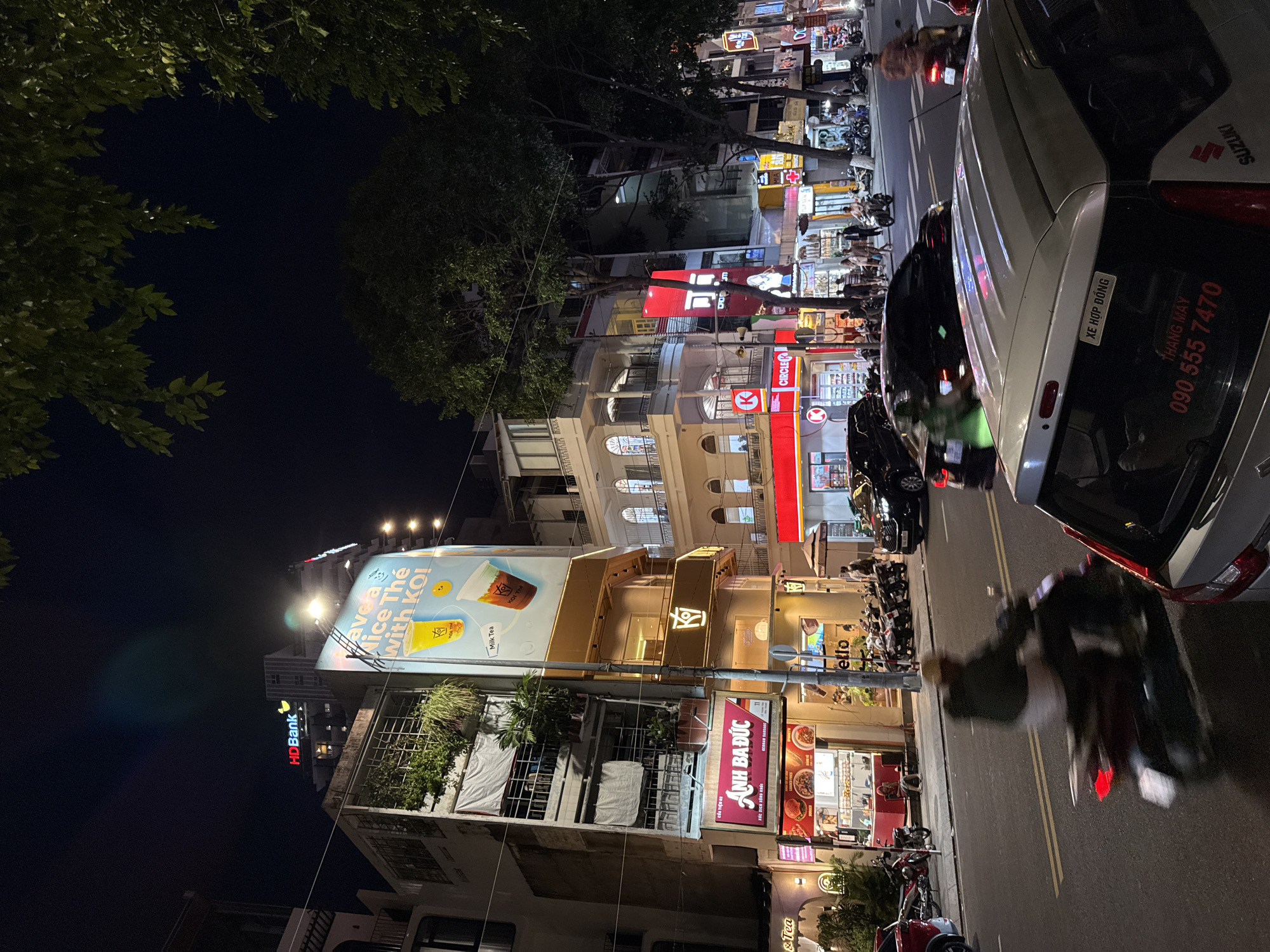 The image depicts a bustling street scene in Nha Trang, Vietnam, at night. The street is lined with multi-story buildings adorned with illuminated signs for various businesses, including "HD Bank" and "KOI Thé." The scene is illuminated by streetlights and the glow of commercial signage, creating a vibrant urban atmosphere.
On the left side of the street, a building features a large billboard advertising "Have a Nice Thé with Koi!" showing images of drinks and a cartoon duck. Below this, a storefront with red and white signage reads "Anh Ba Duc," likely a food establishment, with images of food items displayed. Several motorbikes are parked along the sidewalk, and a few people are visible near the storefronts.
The road is busy with traffic, including several cars and numerous motorbikes. A person is riding a motorbike in the foreground, captured in motion blur, with their headlight and taillight visible. Another motorbike is parked on the side of the road, and a person in a green jacket is riding another motorbike, also with motion blur. A black car is driving in the street, and a silver car is parked in the foreground, partially obscuring the view of the street.
Trees with lush green foliage line the right side of the street, their branches extending into the night sky. The overall impression is a dynamic and lively urban environment, characteristic of a busy commercial district in a Vietnamese city at night.