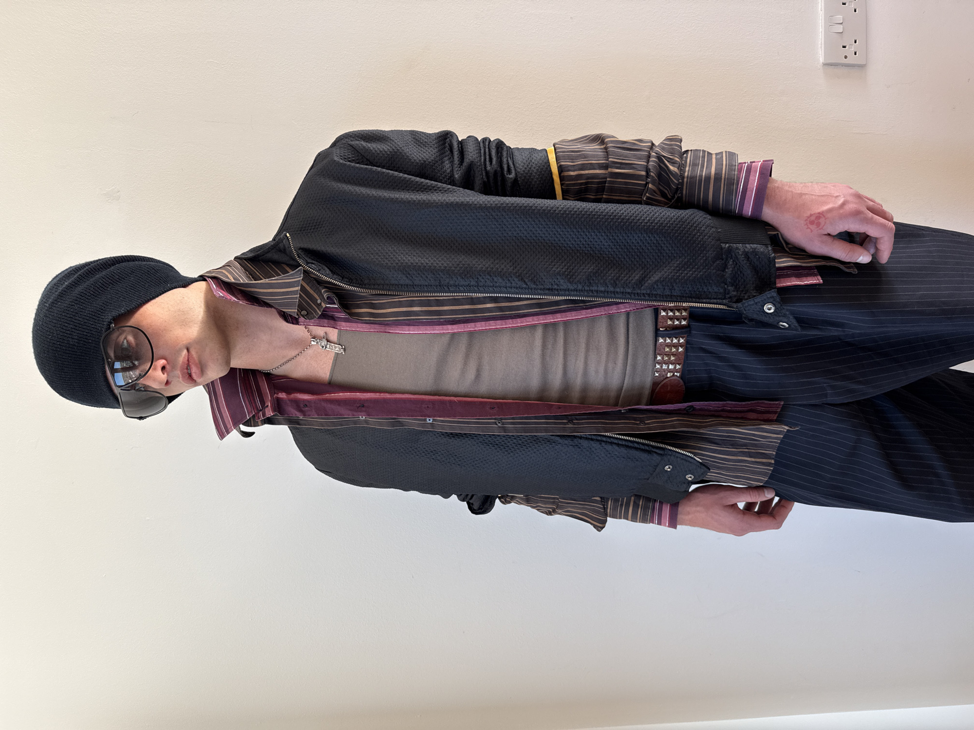 A person is depicted in a full-body shot, standing indoors against a plain, light-colored wall, possibly beige or off-white. The image is oriented vertically, showing the individual from head to approximately mid-shin.
The subject, appearing to be male, is dressed in a layered, stylish outfit. They wear a black beanie and dark sunglasses, which obscure their eyes. A silver necklace with a cross-like pendant is visible around their neck. Underneath a dark, textured, possibly black, jacket with a zipper, a light brownish-tan t-shirt or tank top is worn. Beneath the jacket, a long-sleeved striped shirt in warm tones, featuring brown, gold, and burgundy/purple stripes, is visible, with its cuffs peeking out from under the jacket sleeves. The person wears dark, pinstriped trousers, held up by a brown belt adorned with prominent silver stud or pyramid detailing.
The subject's left hand is visible, resting by their side, and features a small, reddish, circular tattoo on the back of the hand, which appears to be a stylized skull or similar emblem. The person’s facial expression is neutral, and they appear to be posing for the photograph.
In the background, on the right side of the wall, there is a white electrical outlet with two three-pin sockets and two switch buttons. The lighting is soft and even, suggesting an indoor setting with ambient light. No discernible text is visible on the clothing or background, apart from the outline of the pendant. The scene captures a moment of casual posing, likely in a residential or public indoor space.