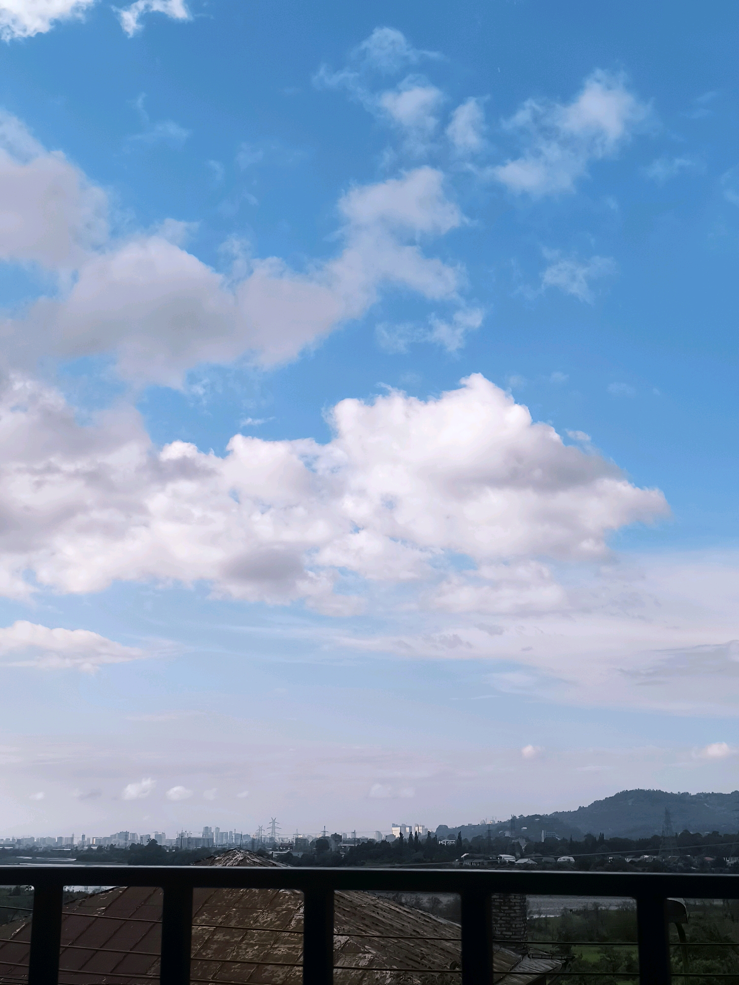 A view from a balcony overlooking a city skyline in Batumi, Georgia, under a bright blue sky with scattered white clouds. The foreground features a dark railing and a rustic, tiled roof. Beyond the railing, a cityscape with numerous buildings, some appearing to be skyscrapers, is visible, stretching towards a distant, tree-covered mountain range. Power lines and other industrial structures are also present in the mid-ground. The overall impression is one of urban development against a natural backdrop.
