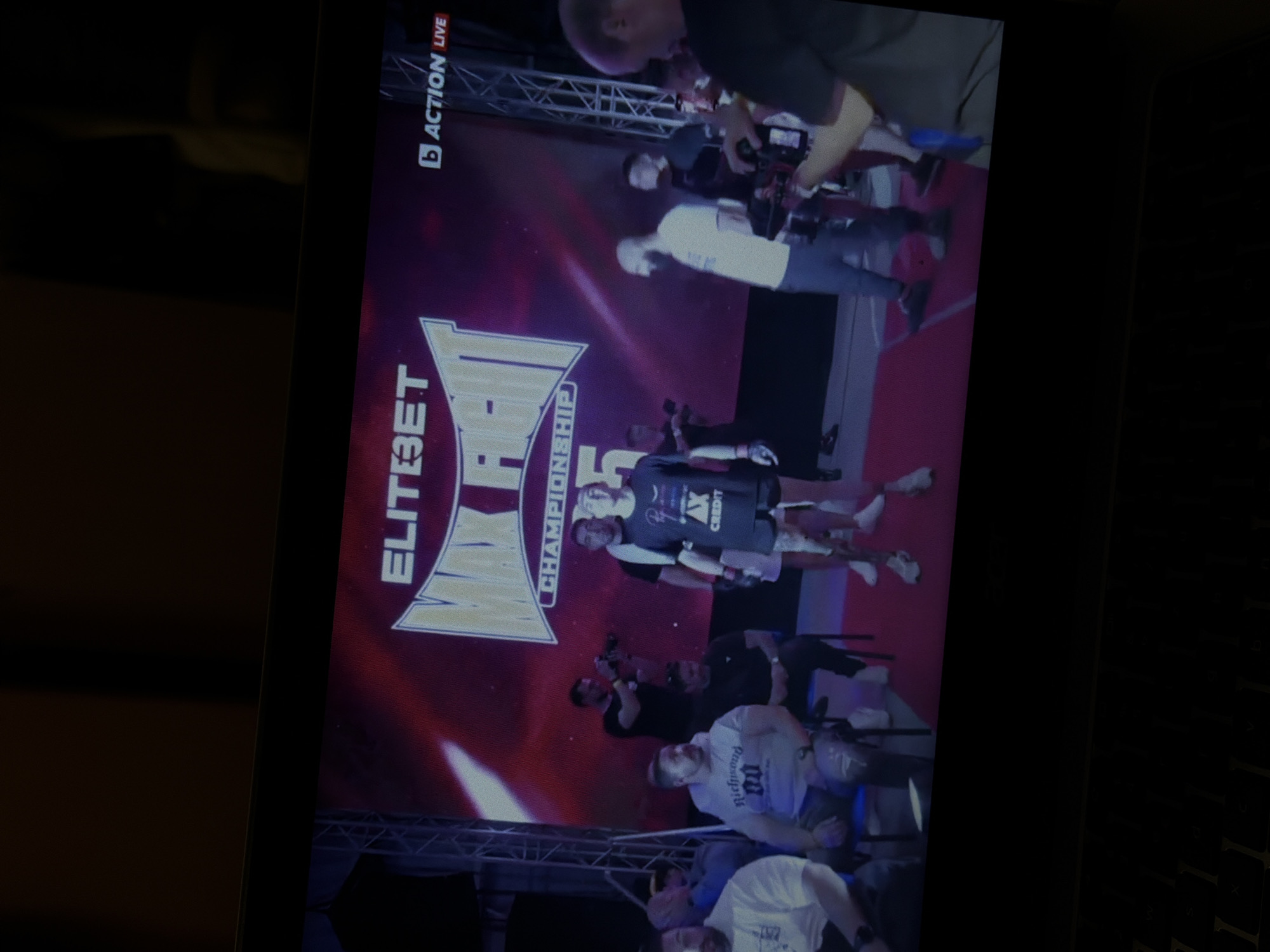 A screen displays a promotional image for ELITEBET MMA Championship, with people visible on the screen. The screen is indoors, likely in a building in Sofia, Bulgaria. Individuals are on and around the screen, some appearing to be filming or taking pictures.