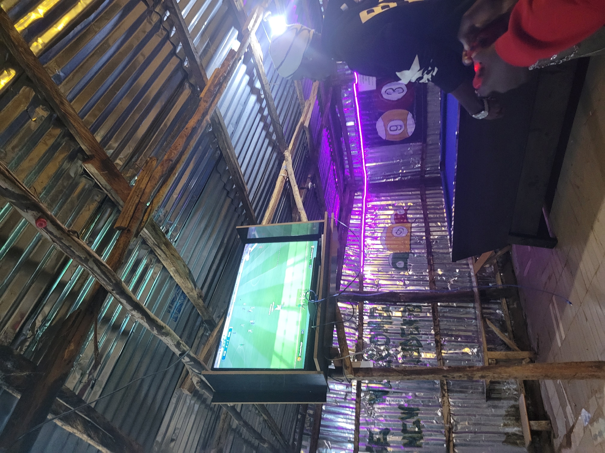 The image depicts an indoor, likely outdoor covered, setting in Durumi, Nigeria. A large television screen displays a soccer game. A person wearing a white cap and a dark shirt is visible on the right, with their back mostly turned to the camera. They appear to be holding a red gaming controller. In the foreground, another person's hands are visible, also holding a red controller. A pool table with a blue surface is situated in the mid-ground. The environment is rustic, with a corrugated metal roof supported by wooden beams and corrugated metal walls. Purple neon lights are installed along the walls. Some pool balls with numbers are visible on a display behind the pool table. Graffiti is present on the metal walls. The flooring appears to be wood or concrete.