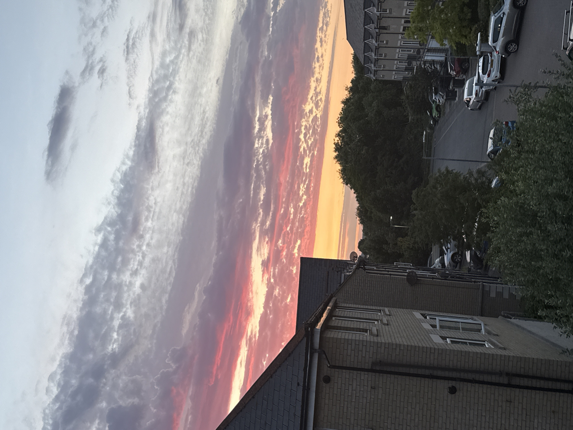 A view from a residential building in Great Yarmouth, United Kingdom, captures a colorful sunset over a street lined with parked cars and trees. The sky is filled with dramatic clouds, a mix of white, grey, and vibrant pinks and oranges from the setting sun. In the foreground, the side of a brick building with dark grey roofing is visible. Below, a paved street curves through the scene, with several cars parked along its sides. These include a silver SUV, a white hatchback, a blue hatchback, and other vehicles further down the street. Dense green trees form a backdrop, with some foliage extending into the lower portion of the frame. The scene depicts a typical residential street during twilight.