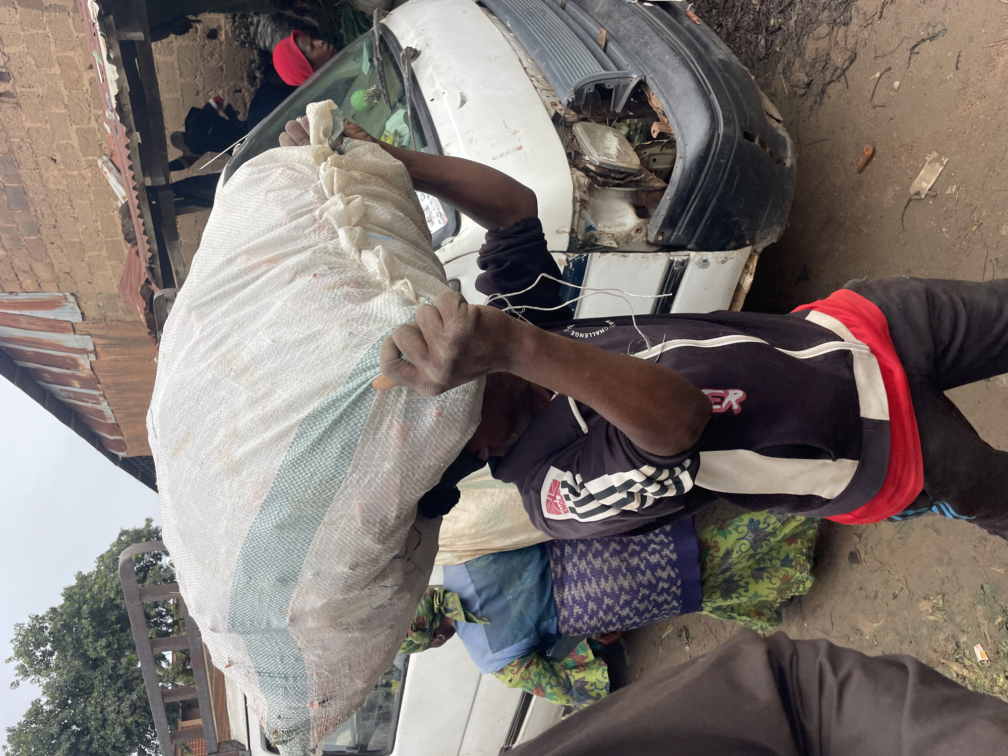 The image captures a scene in Gura Top, Nigeria, likely during the daytime given the bright lighting. A person is depicted from the chest down, actively carrying a large, white woven bag filled with what appears to be goods or refuse. The bag is supported by the individual's shoulders and is partially obscuring their face. They are wearing dark clothing with red and white stripes on the sleeves, and a red cloth is visible around their neck.
Behind the person, the front of a white car is visible, its front end significantly damaged, with broken headlights and exposed engine components, suggesting it may be a derelict vehicle or undergoing repair. The surrounding environment appears to be an outdoor, possibly informal, setting. A brick wall with a corrugated iron roof is visible to the left. The ground is dirt and uneven.
Other individuals can be seen in the background, including one wearing a red head covering, also appearing to be involved in some activity related to the car or the cargo. Colorful patterned fabrics are also visible, possibly being transported or stored. The overall impression is one of manual labor or salvage work in a somewhat cluttered, possibly rural or semi-urban, environment. The mood is neutral, focusing on the task at hand. No discernible text is present in the image.