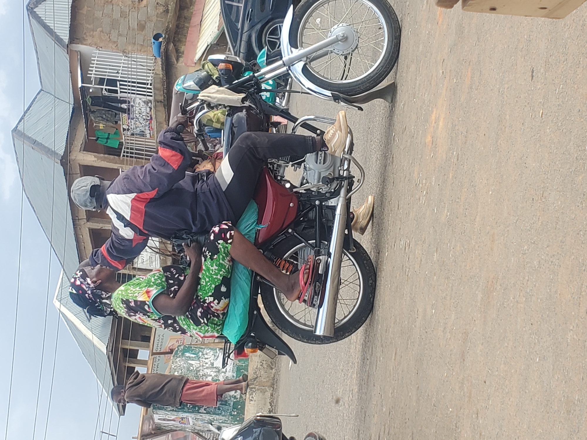 The image shows two people on a motorcycle in Gwong, Nigeria. The person in front is a man wearing a dark jacket with red and white stripes on the sleeves and a grey helmet. He is riding the motorcycle. Behind him, a woman wearing a colorful patterned dress and a headscarf is seated. She has a light blue cloth draped over her. Both are facing forward, with the man appearing to be in motion and the woman looking slightly to her side.
The motorcycle is dark red with chrome accents and a silver wheel. The scene appears to be a street in a town. In the background, there are buildings with corrugated metal roofs, some shops with displays of clothing and goods behind metal grates, and a few other motorcycles. A young boy wearing a brown hooded sweatshirt and orange shorts is visible standing in the middle ground, looking towards the motorcycle.
The lighting suggests it is daytime, likely under a clear sky. The overall atmosphere is casual and appears to be a normal day on a busy street. There is no visible text in the image.