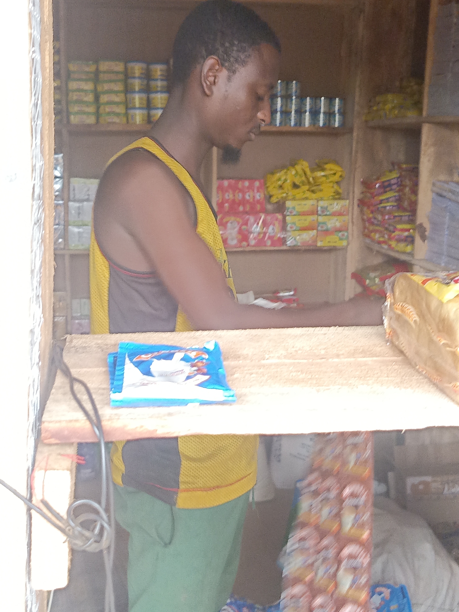 The image captures a man standing behind a wooden counter within a small shop in Akwanga, Nigeria. The shop's shelves are stocked with various goods, including canned goods, packaged foods, and other items. The man is wearing a yellow and black tank top and green pants. He appears to be waiting or attending to customers. A package of blue and white goods is on the counter in front of him. The lighting suggests it's daytime, and the environment is typical of a small retail establishment in a developing urban area.