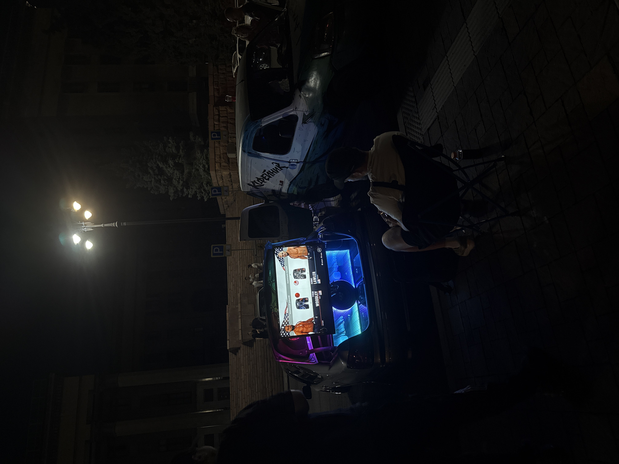 The image shows a dimly lit outdoor scene at night, likely in Almaty, Kazakhstan, as suggested by the provided context. A white van with a cartoonish mural and the Cyrillic word "Кофеман" (Kofeman, meaning coffee lover) is parked on a street with a brick sidewalk. The van is outfitted with a large, illuminated screen displaying what appears to be a combat sports event, featuring images of fighters like Tyson Woodley and Lawler. The screen is surrounded by vibrant blue and pink LED lights.
Several people are visible around the van, some standing and observing the screen, while one person is leaning over the screen, possibly interacting with the equipment. A street lamp with three bright lights illuminates a portion of the scene. In the background, there are buildings and trees, but they are mostly obscured by darkness. Two parking signs with the letter "P" are visible on poles near the van. The overall atmosphere suggests a public gathering or event, possibly a viewing party or a mobile entertainment setup, taking place after dark. The lighting and the focus on the illuminated screen create a dynamic and somewhat festive mood, despite the darkness.