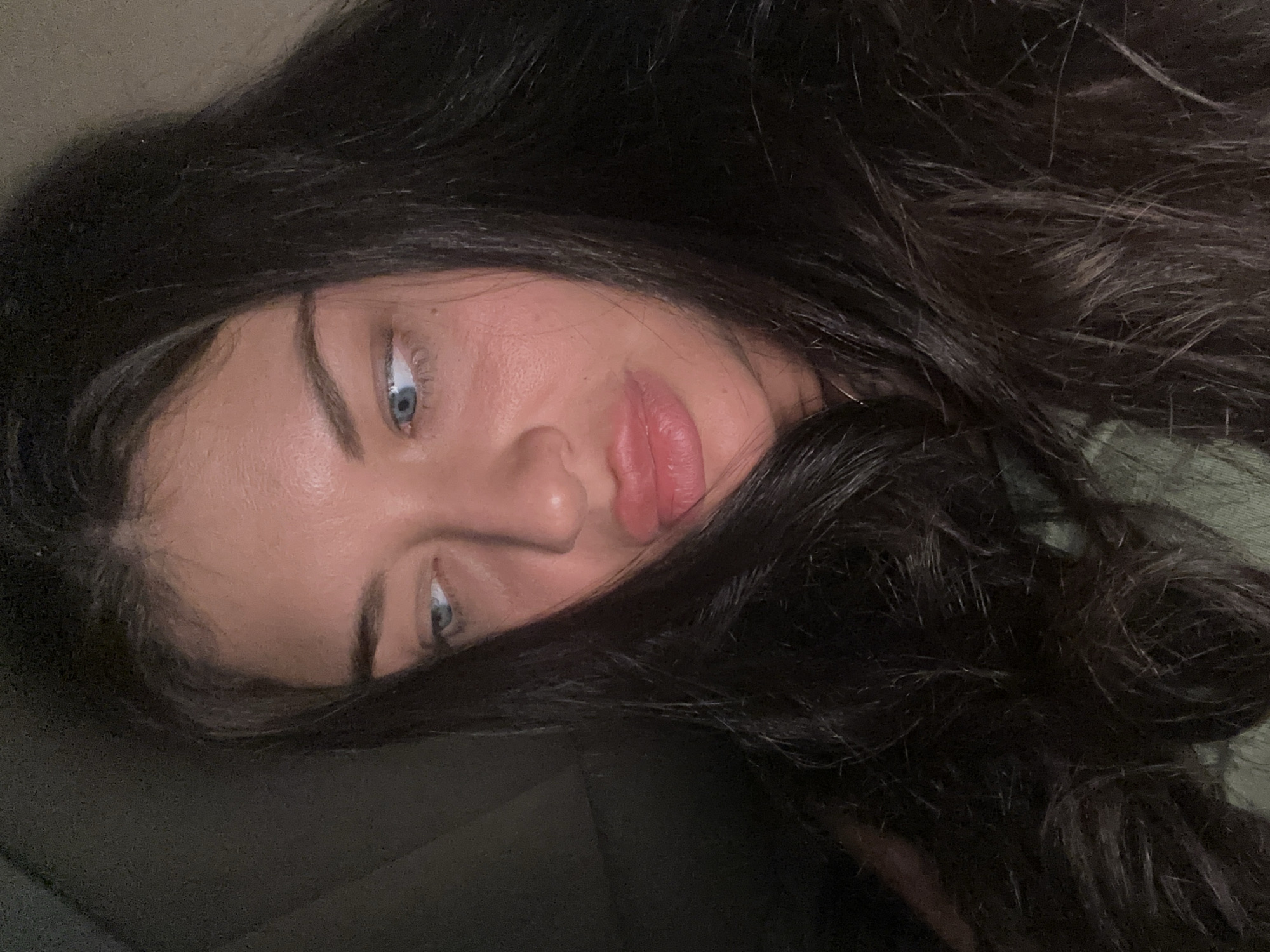 A close-up portrait, likely a selfie, features a woman in a reclined or horizontal pose, taken in Khimki, Russia. Her face is tilted, with her light blue eyes looking subtly upwards or towards the viewer. She has dark, long, wavy hair that frames her face, flowing across much of the frame and creating a dark backdrop. Her lips are full and appear to have a natural pinkish hue. Her skin is clear with a natural complexion, showing a slight flush on the cheeks. A glimpse of a light green garment is visible on the right side of the image. The overall lighting is dim, suggesting an indoor setting with low light, possibly during the evening or night. The background is indistinct and dark, emphasizing the subject. Her expression is neutral and calm. There is no visible text.