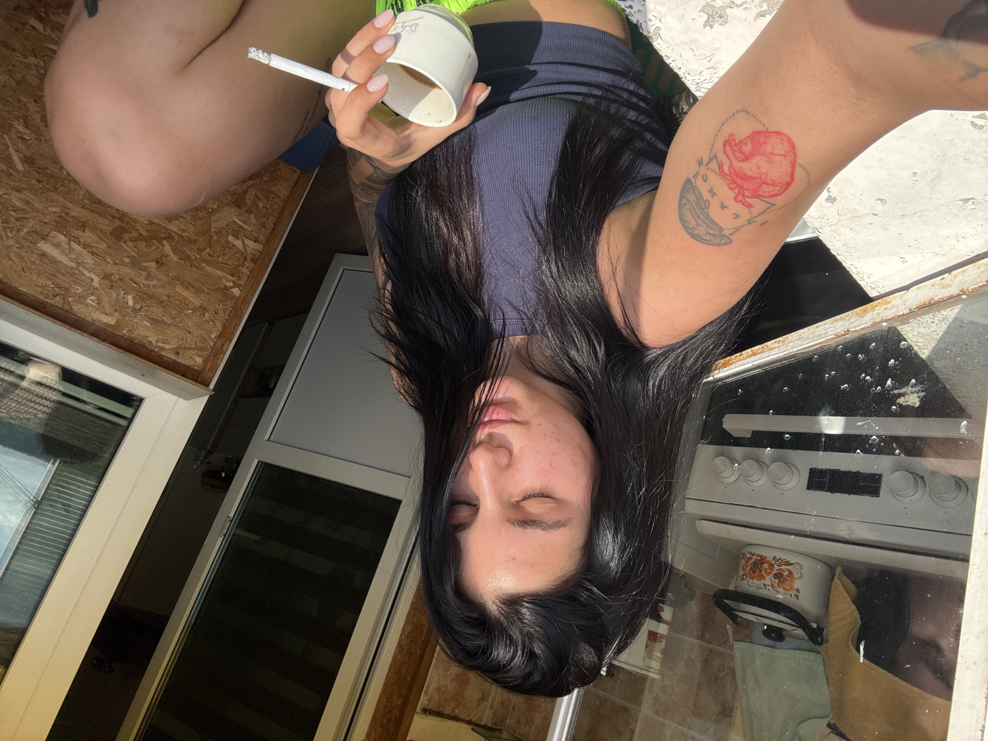 A young woman with dark hair is shown in an upside-down, high-angle perspective. She holds a cigarette and a cup. She has a tattoo of a red rose or heart on her arm. The background includes a window, an interior with what appears to be a stove and other household items. The scene appears to be indoors. The location of Veliko Tarnovo, Bulgaria, is not evident in the image.