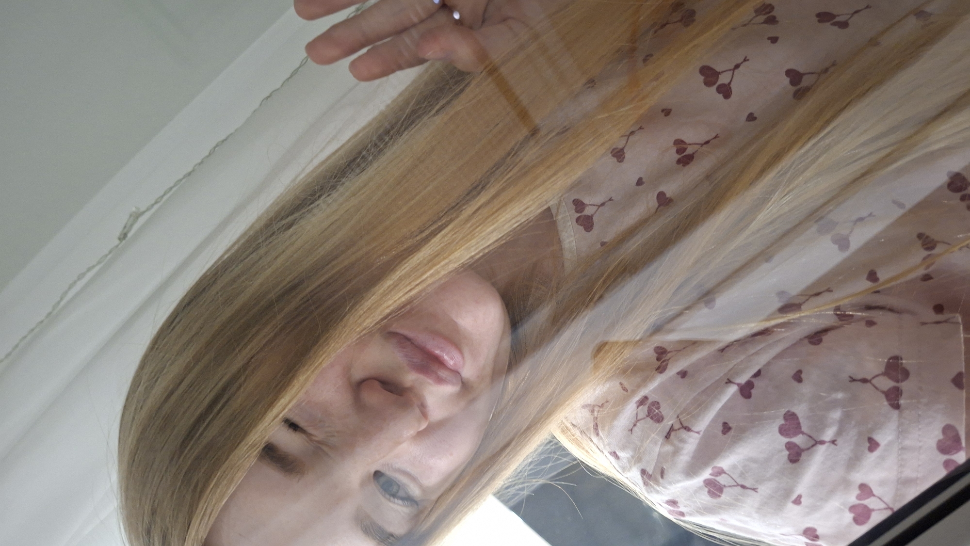 A blonde woman is looking down at the viewer. She is wearing a pink shirt with a red heart pattern. Her hair is long and straight. Her right hand is near the top of the frame. The background includes a white curtain. The location is not discernible from the image.