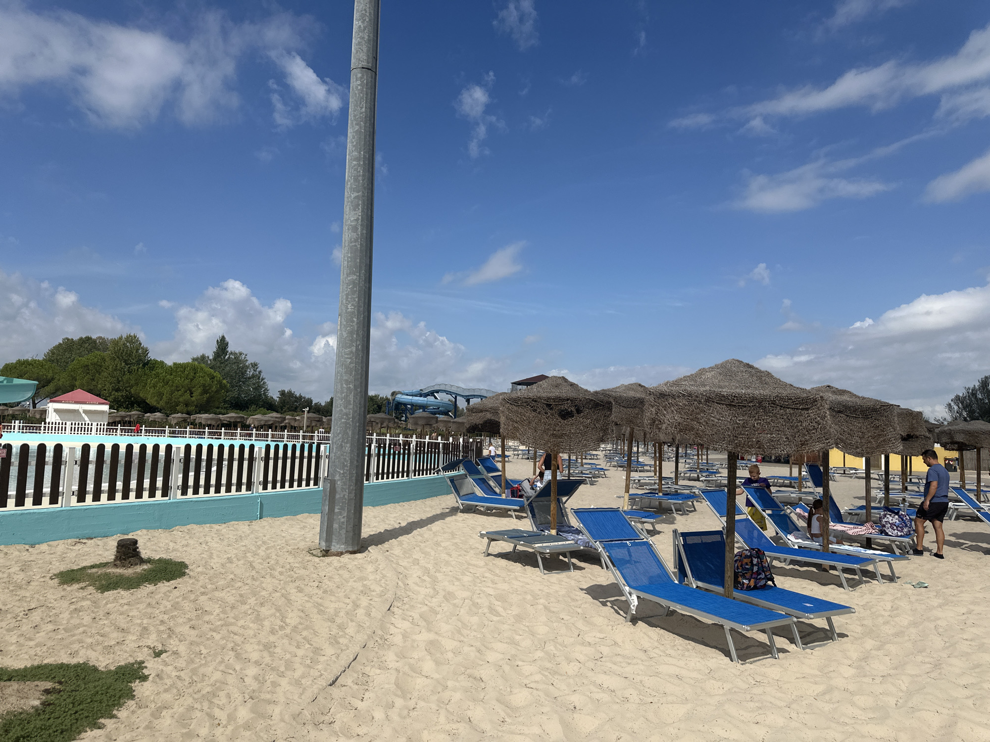 The image depicts a sandy beach with numerous blue lounge chairs and straw umbrellas, suggesting a resort or water park environment. In the background, a swimming pool is visible, bordered by a fence and a bright turquoise wall. Beyond the pool, a red-roofed building with "GELATO" signage can be seen, along with lush green trees. Further in the distance, blue water slides characteristic of a water park are present. A tall, silver metallic pole stands prominently in the foreground on the left side of the image. Several people are scattered among the lounge chairs, some sitting and others standing. The sky is a clear blue with scattered white clouds. The scene appears to be a sunny day at a leisure facility.