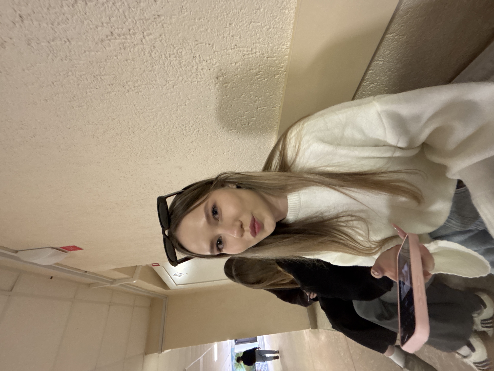 A young woman is taking a selfie in a building in Chaikovsky, Russia. She has long blonde hair and is wearing a white sweater. Sunglasses are perched on her head. She is holding a pink smartphone in her hand. Another person is partially visible in the background, wearing a black jacket. A man is walking away in the far distance. The location appears to be a hallway or corridor with beige walls and a white ceiling.