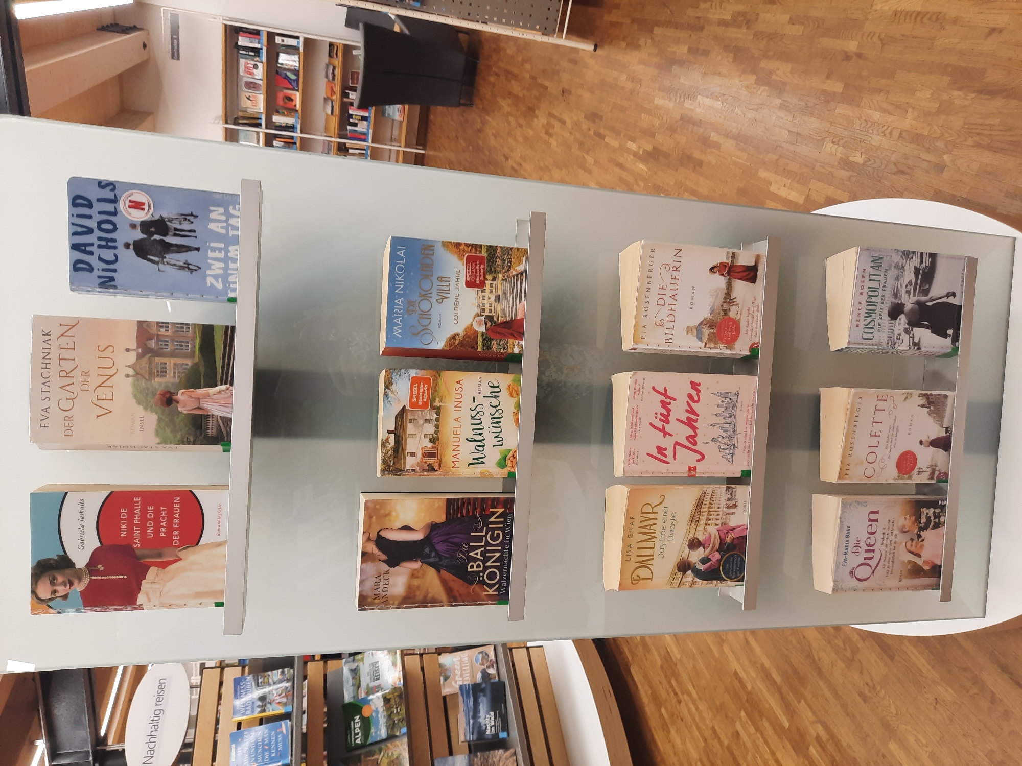 The image shows a retail display of books in a store, likely a bookstore or department store. The display is set against a light blue wall, with white shelves holding various titles. The books are arranged in rows, with some standing upright and others leaning against each other. The spines of the books are visible, showing titles, authors, and some cover art.
In the top row, on the left, is a book titled "Gabriela Jaskulla NIKI DE SAINT PHALLE UND DIE PRACHT DER FRAUEN". Next to it is a book by Eva Stachniak titled "DER GARTEN DER VENUS". To the right of that is a book by David Nicholls titled "ZWEI AN EINEM TAG".
The middle row features books such as "DIE BALL KÖNIGIN" by Mara Andeck, "Walnuss-Wünsche" by Manuela Inusa, and "DIE Schokoladen VILLA GOLDENE JAHRE" by Maria Nikolai.
The bottom row displays "Dallmayr Das Erbe Einer Dynastie" by Lisa Graf, "In Fünf Jahren" by Eva Rosenberger, and "DIE BILDHAUERIN" by Pia Rosenberger. Further down, on the bottom shelf, are "Die Queen" by Eva-Maria Bart, "COLETTE" by Pia Rosenberger, and "COSMOPOLITAN DIE ZEIT DER FRAUEN" by Renee Rosen.
To the left of the book display, there is a sign that reads "Nachhaltig reisen". Below this sign, on a lower shelf, are more books, some with travel-related titles and images, such as "ALPEN" and "Green Travelling".
The environment appears to be indoors, with wooden flooring visible at the bottom of the image. In the background, to the right, is a glimpse of a shelving unit filled with more books, and a dark armchair. The lighting is bright and even, illuminating the book display. The context of Rosenheim, Germany, is not directly evident from the image content itself, but the book titles are in German, suggesting a German-speaking region. The presence of the specific book titles does not inherently place the scene within Rosenheim unless further information is provided.