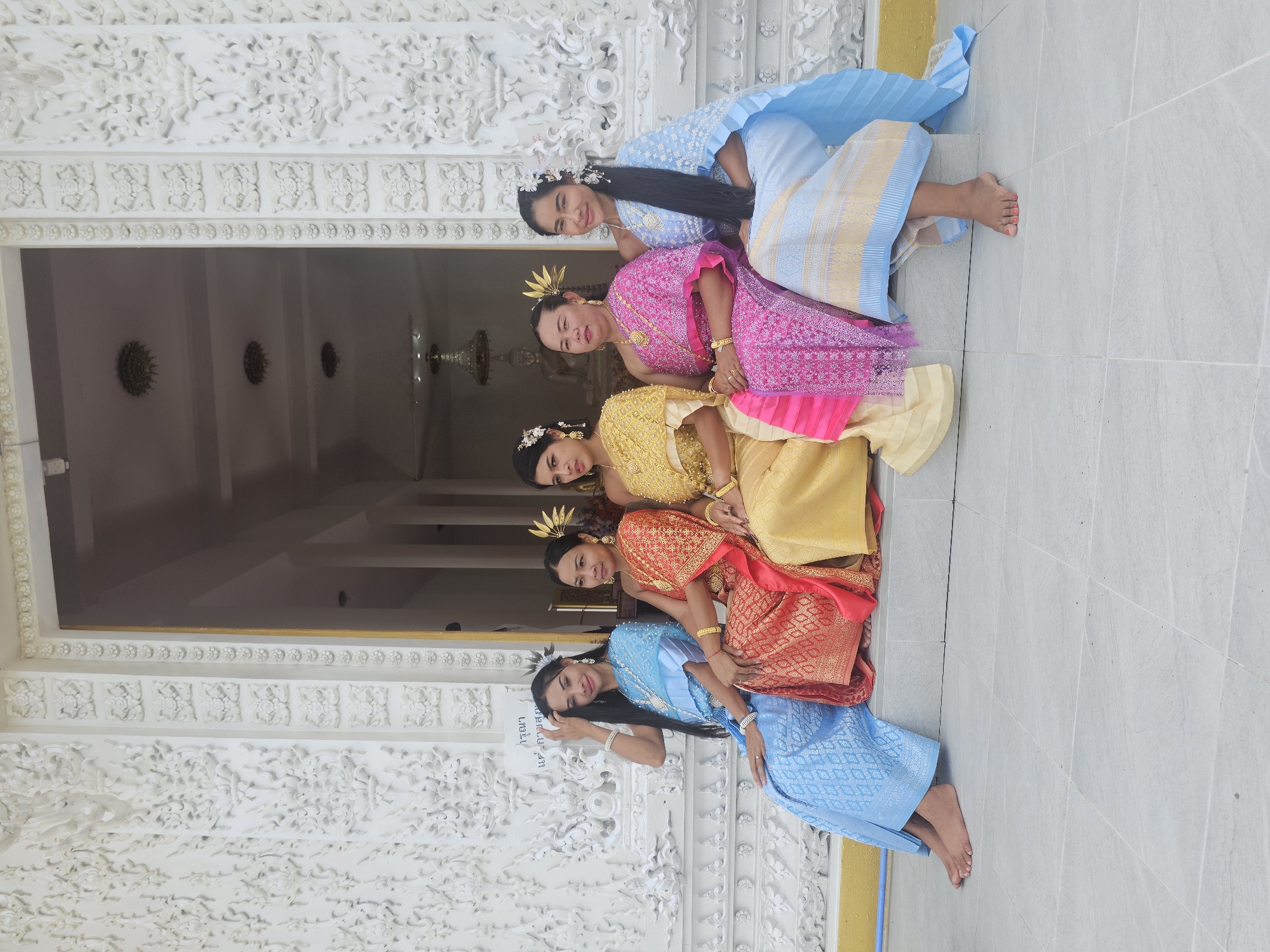 Five women are seated on a step in front of a white ornate building, likely a temple, in Thep Krasatti, Thailand. All five women are wearing traditional Thai clothing. From left to right, the first woman wears a light blue silk outfit. The second woman is dressed in a red and gold outfit. The third woman wears a golden yellow outfit with pink accents. The fourth woman is in a pink and gold outfit. The fifth woman on the far right wears a light blue outfit with gold accents. They are all adorned with jewelry and some have flowers in their hair. The building behind them features intricate white carvings and a large entryway. The ground in front of them is tiled. The overall scene depicts a cultural gathering or celebration.