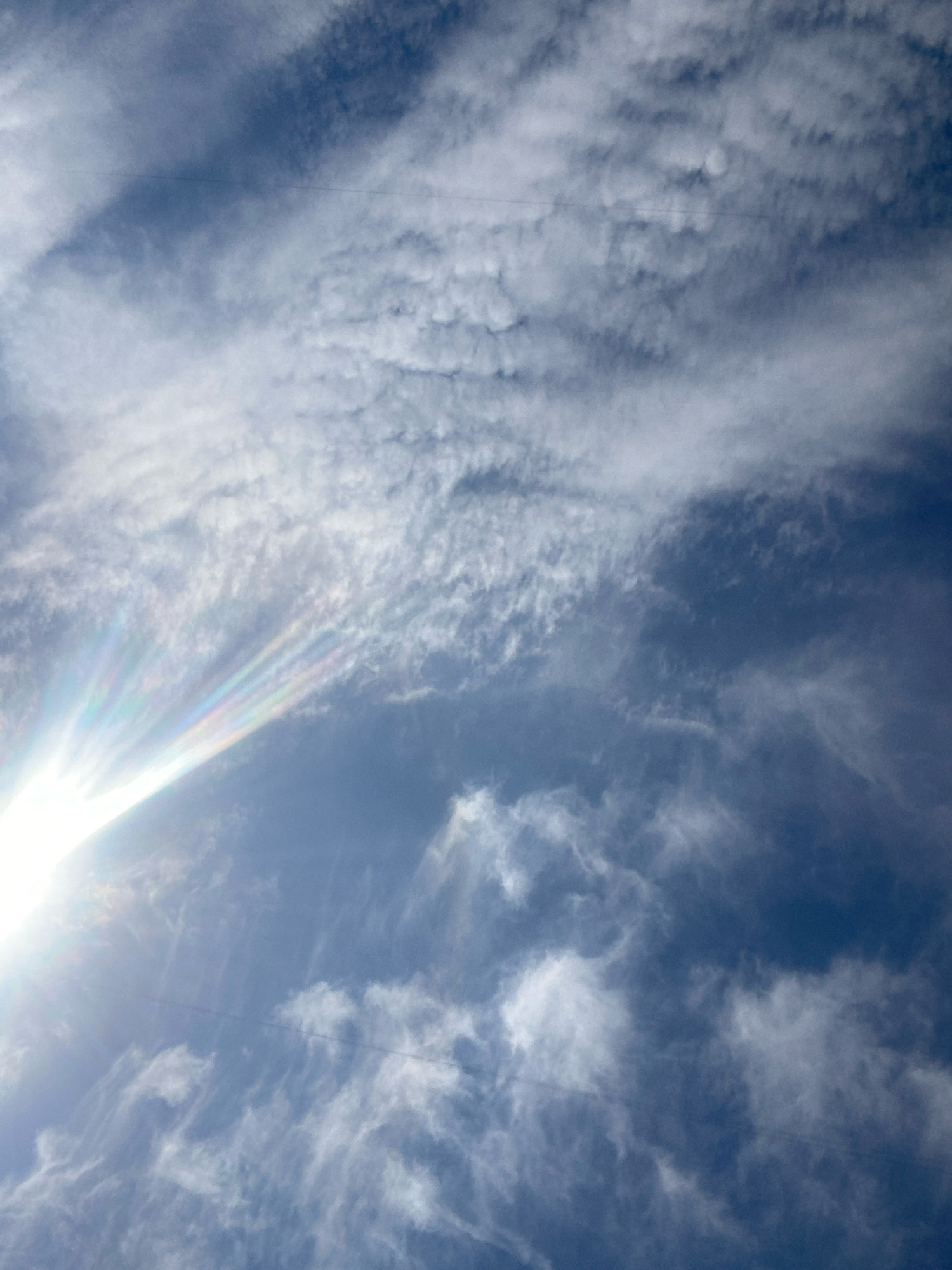 A clear blue sky is visible with wispy, white clouds. The sun, positioned in the lower left quadrant, casts a bright glare and a faint rainbow-colored halo effect onto the clouds. A thin, dark horizontal line, possibly a power line, is visible across the upper portion of the image. The scene is an outdoor environment, likely Chornomorsk, Ukraine, given the context. No people or specific landmarks are discernible.