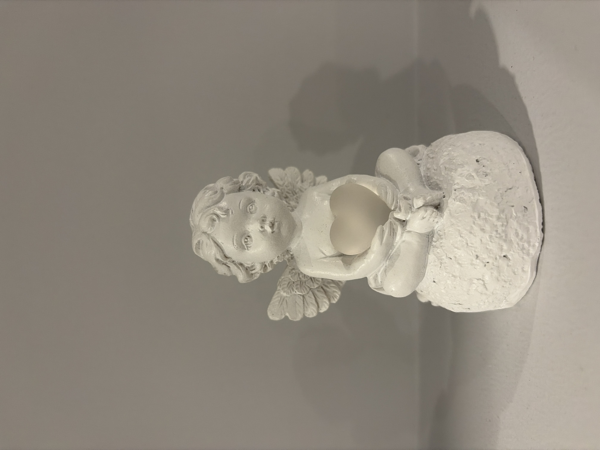 A white cherub figurine sits on a textured base, holding a heart-shaped object. The figurine is positioned centrally in the frame, with its wings spread slightly. The background is a plain, light gray wall. The figurine rests on a white surface, possibly a shelf, which extends to the bottom of the image. A shadow of the figurine is cast on the wall to its right. The image does not contain information about the city of Podgumer, Bulgaria.
