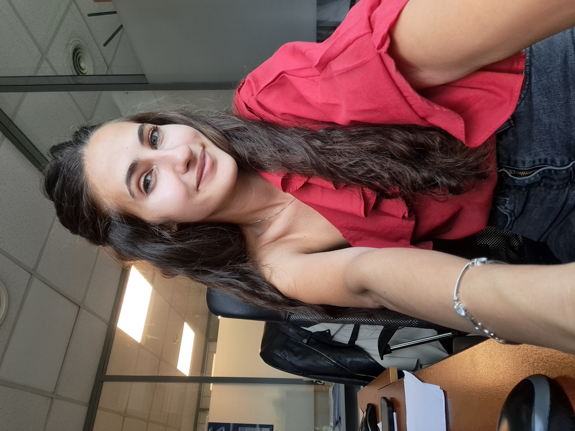 A woman with long, dark brown hair and fair skin is seated in an office setting. She wears a red blouse and dark denim shorts with a visible zipper. She is smiling slightly. On her wrist, she has a silver bracelet with small, light-colored stones. The office has a drop ceiling with circular vents. Behind her, there is a window, and a black leather jacket hangs on the back of her chair. A desk is visible in the foreground, cluttered with papers and office supplies. It is not possible to confirm from the image if the location is in Sofia, Bulgaria.