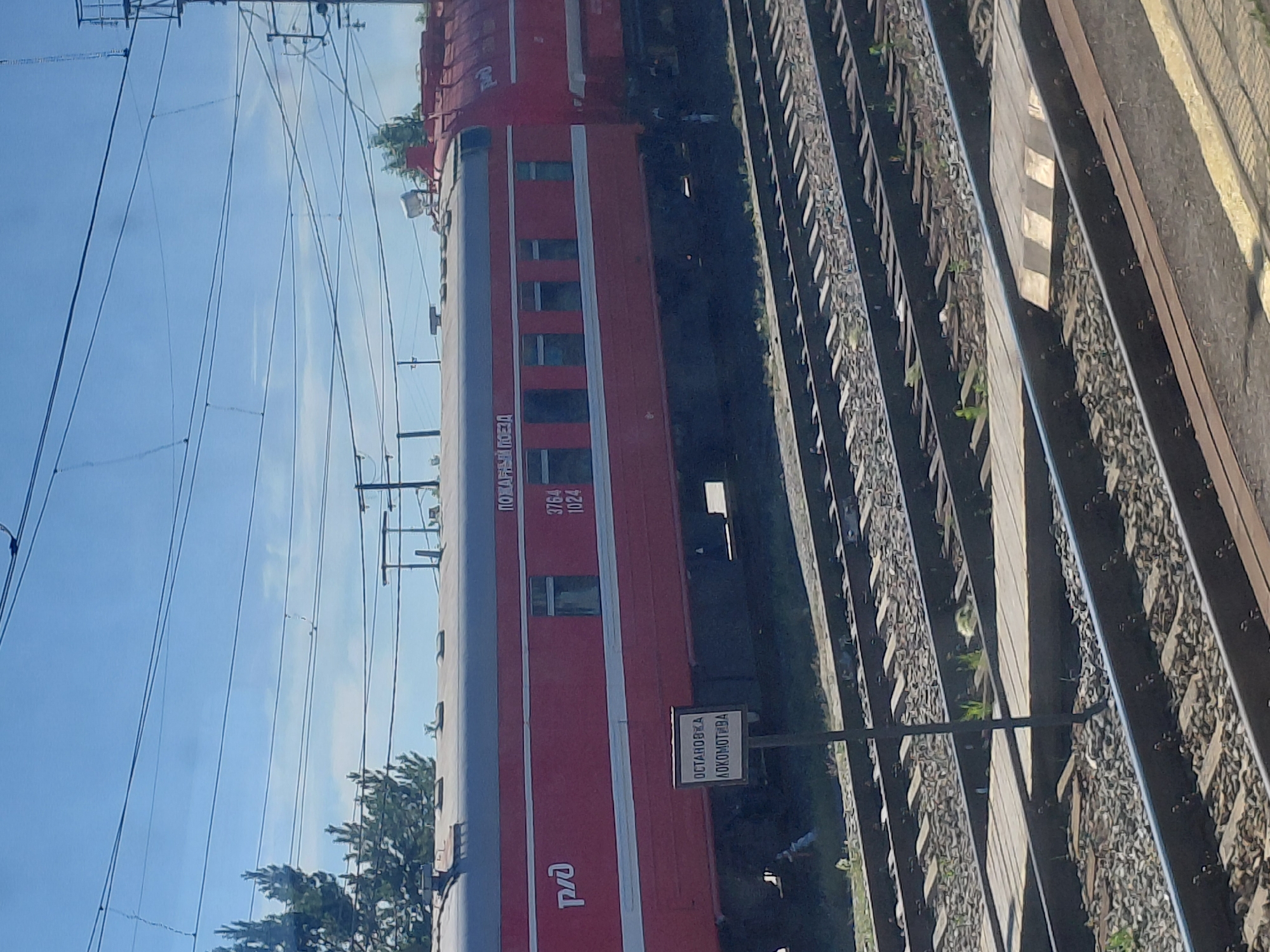 The image shows a red train car with "РЖД" branding on its side, indicating it belongs to Russian Railways. The train car has windows and is positioned on railroad tracks. A signpost with the text "ОСТАНОВКА ЛОКОМОТИВА" (Locomotive Stop) is visible near the train. In the background, there are overhead power lines and trees. The scene appears to be a railway station in the city of Chulym, Russia. The sky is blue with some clouds, suggesting it is daytime. There are no people or specific actions depicted in the image.