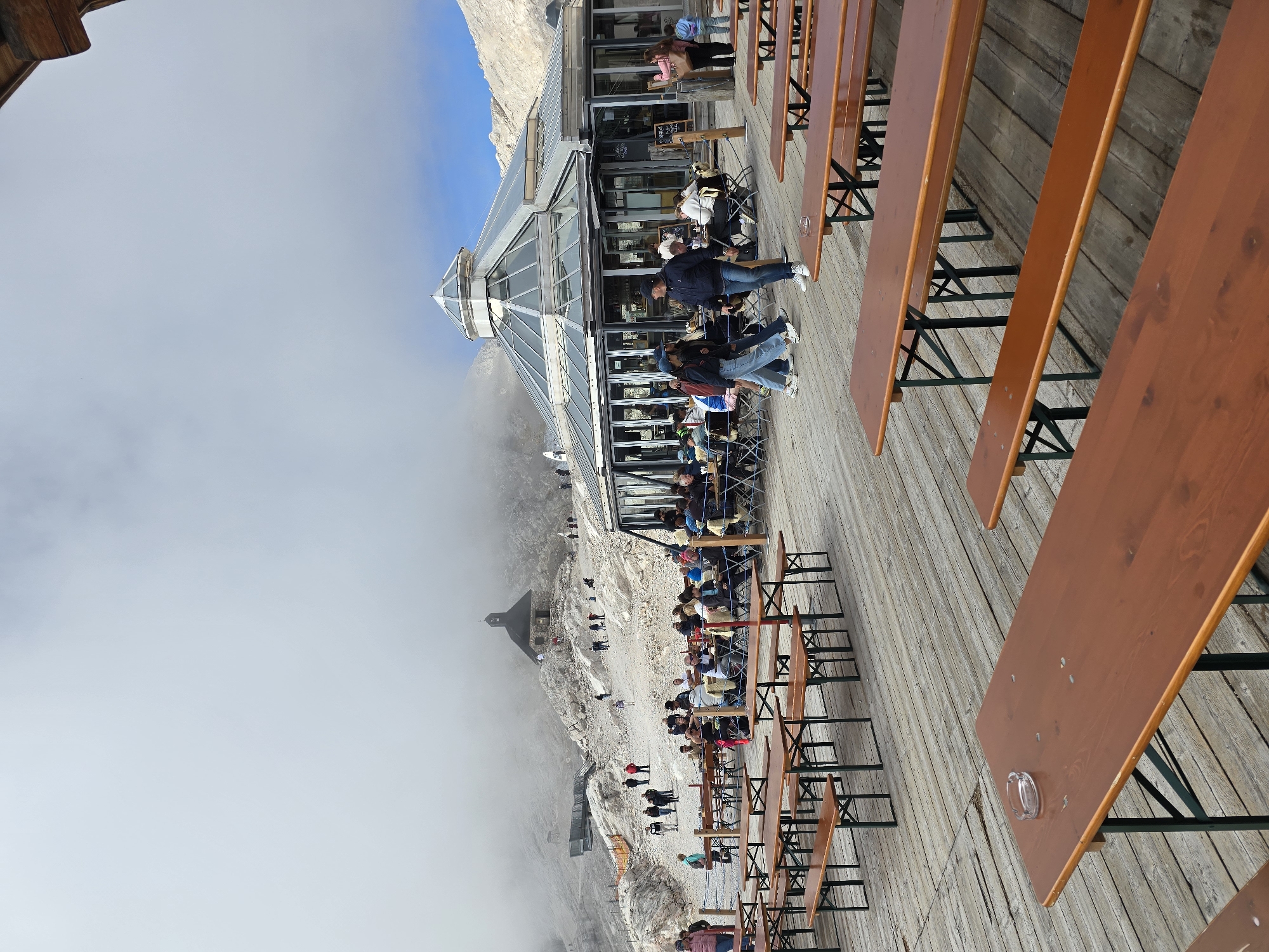 The image depicts an outdoor seating area of a restaurant or beer garden situated at a high altitude, likely in the Bavarian Alps near Garmisch-Partenkirchen, Germany. In the foreground, numerous wooden picnic tables and benches are arranged on a wooden deck. In the midground, a modern glass-domed building, which appears to be the restaurant, is visible, with patrons seated at tables both inside and outside. Further in the background, a small, historic church is perched on a rocky, mountainous terrain. The scene is partially obscured by a layer of thick fog or clouds, giving a sense of elevation and a potentially cool, misty atmosphere. Several individuals are present, walking or seated, enjoying the mountainous landscape.
