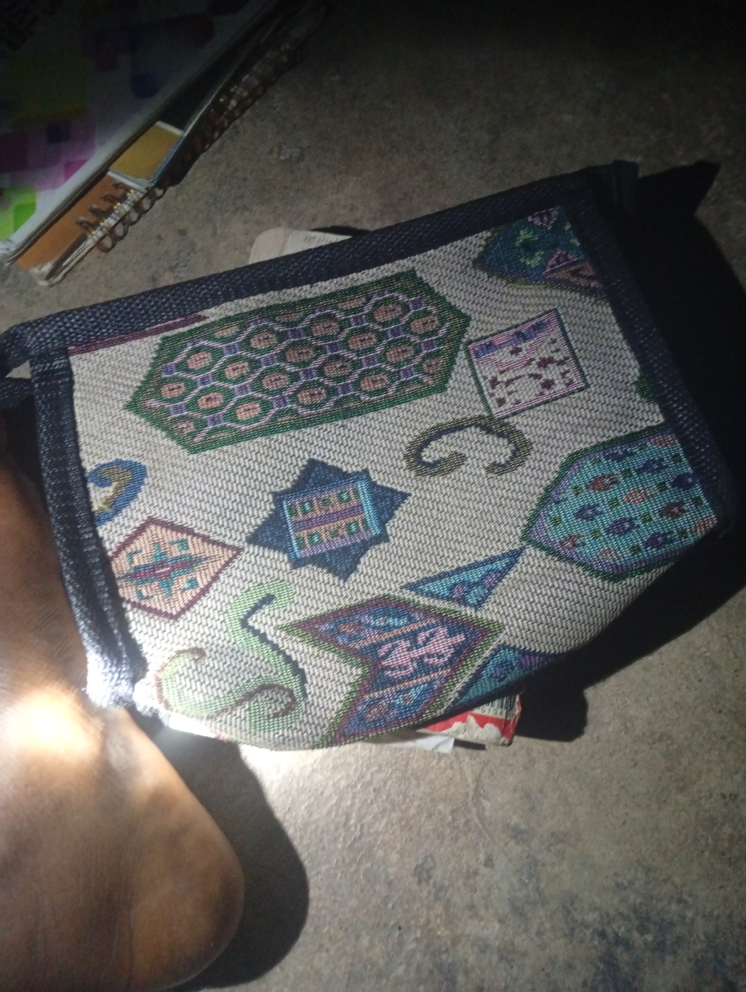 The image shows a close-up view of a woven fabric with geometric patterns in various colors including green, purple, blue, and pink. The fabric appears to be a part of a bag or a mat, with a dark blue border. A person's leg and foot are visible on the left side of the image, casting a shadow on the textured ground. In the background, a spiral-bound notebook is partially visible, with its pages fanned out. The lighting suggests an indoor or dimly lit outdoor environment, possibly at night. The scene is set on a concrete or stone-like surface. The image does not provide enough information to determine its specific location within Jalingo, Nigeria.