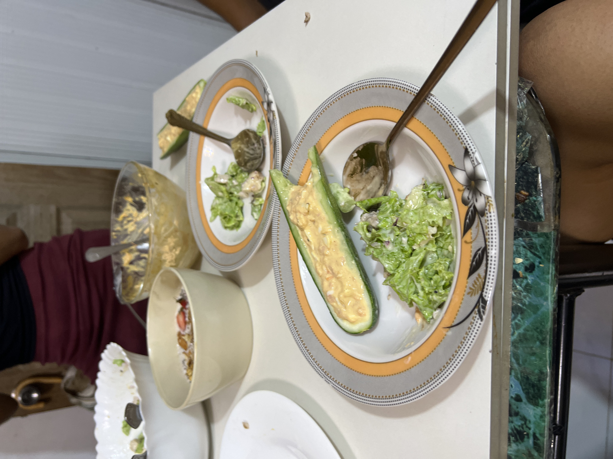 The image depicts a table set for a meal, likely after consumption, with remnants of food and dining utensils. In the foreground, a plate holds a half cucumber stuffed with a creamy yellow mixture and a portion of green salad. A spoon rests on the plate. Another plate with salad and a spoon is visible behind it. In the background, a large bowl contains a creamy substance, and another bowl holds cereal with fruit. A person's torso, wearing a maroon skirt and black top, is partially visible on the left. The setting appears to be indoors, possibly a dining area, with a white table and a glimpse of a green marble-patterned surface below. The overall impression is of a meal that has been eaten, with the food remnants indicating various dishes. The description does not provide information about the specific location in Davao City, Philippines.