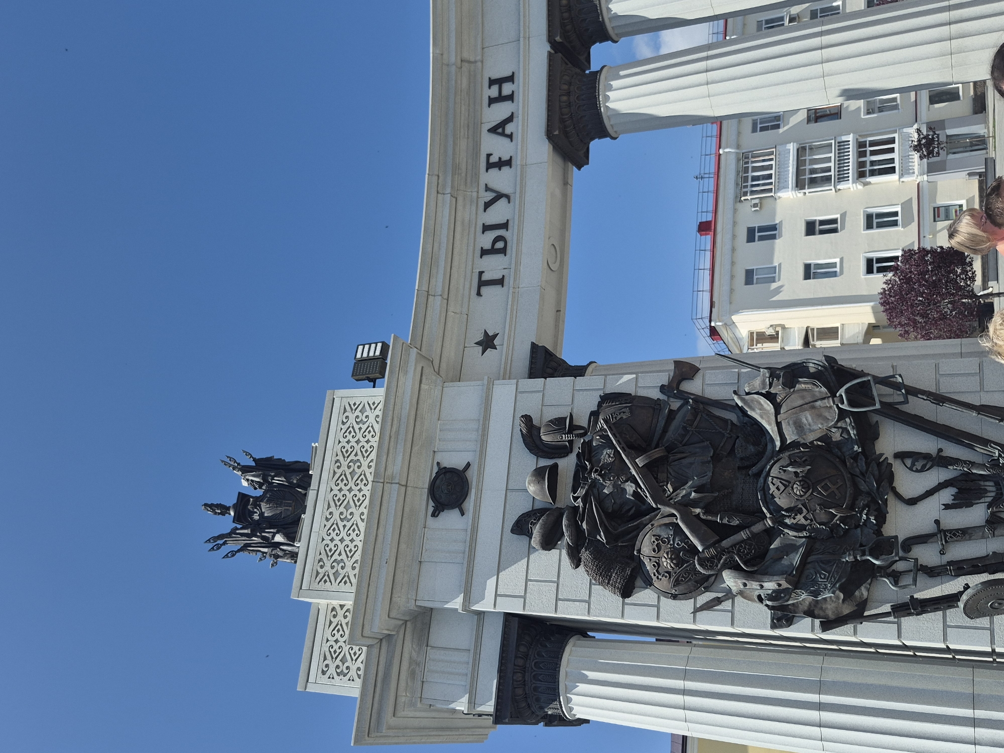 The image shows a view of a triumphal arch in Ufa, Russia, with an inscription that appears to be "ТЫУГАН". The arch is decorated with a large bronze relief depicting several figures in historical armor, shields, and weapons. Above the relief, on the left side of the arch, is a crest with several figures. The arch is supported by large, fluted white columns. In the background, a light-colored, multi-story building with a red roof is visible, along with a small tree. The sky is clear and blue. Several people are partially visible in the foreground at the bottom of the image, suggesting a public space.