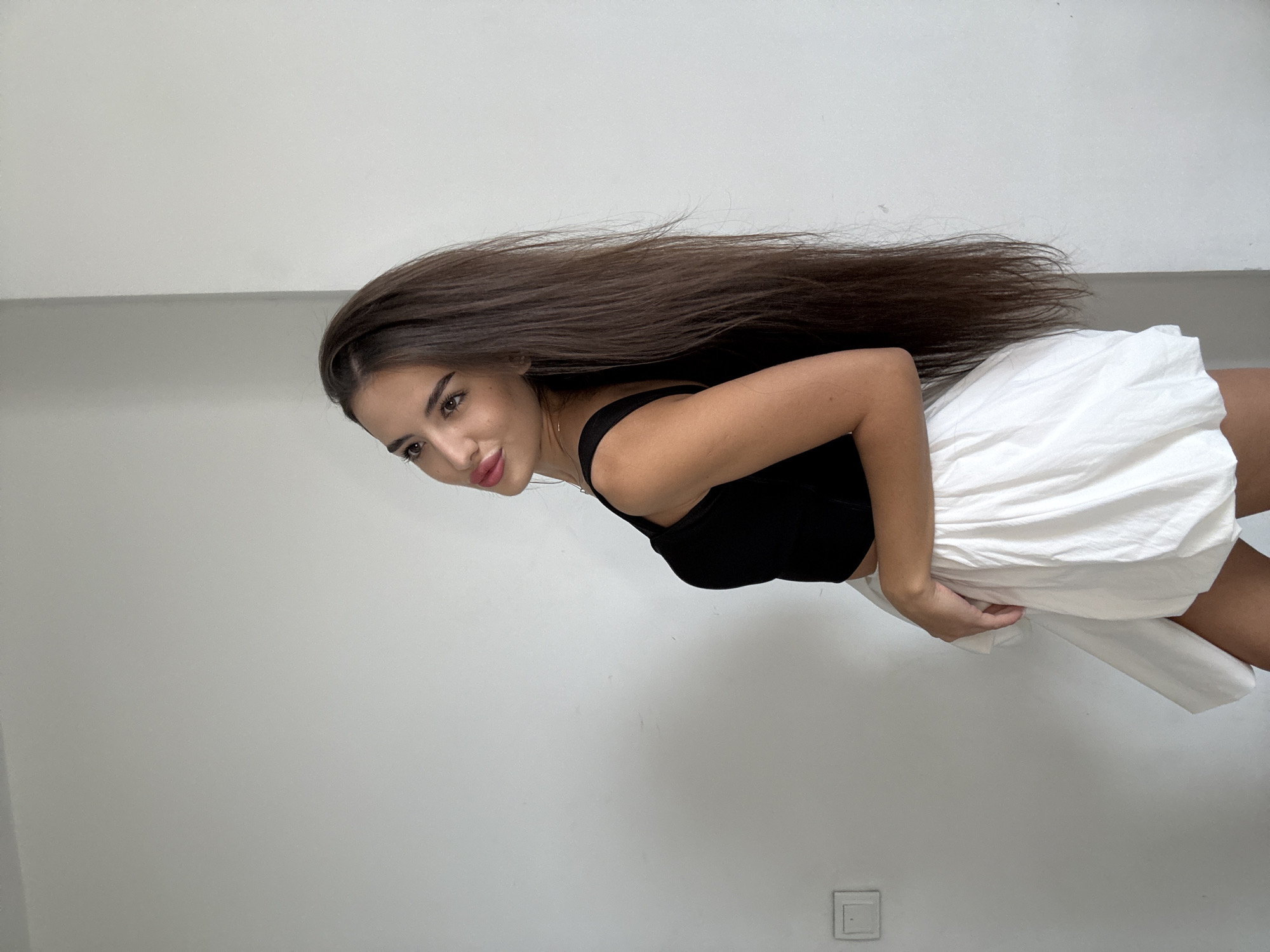 A young woman with long, dark brown hair and light skin is shown from the side, looking over her shoulder at the camera. She is wearing a black tank top and a white, puffy skirt. She has on-trend makeup, including winged eyeliner and pink lipstick. The background is a plain white wall with a light switch on the left side. The image was taken indoors in Chișinău, Moldova.