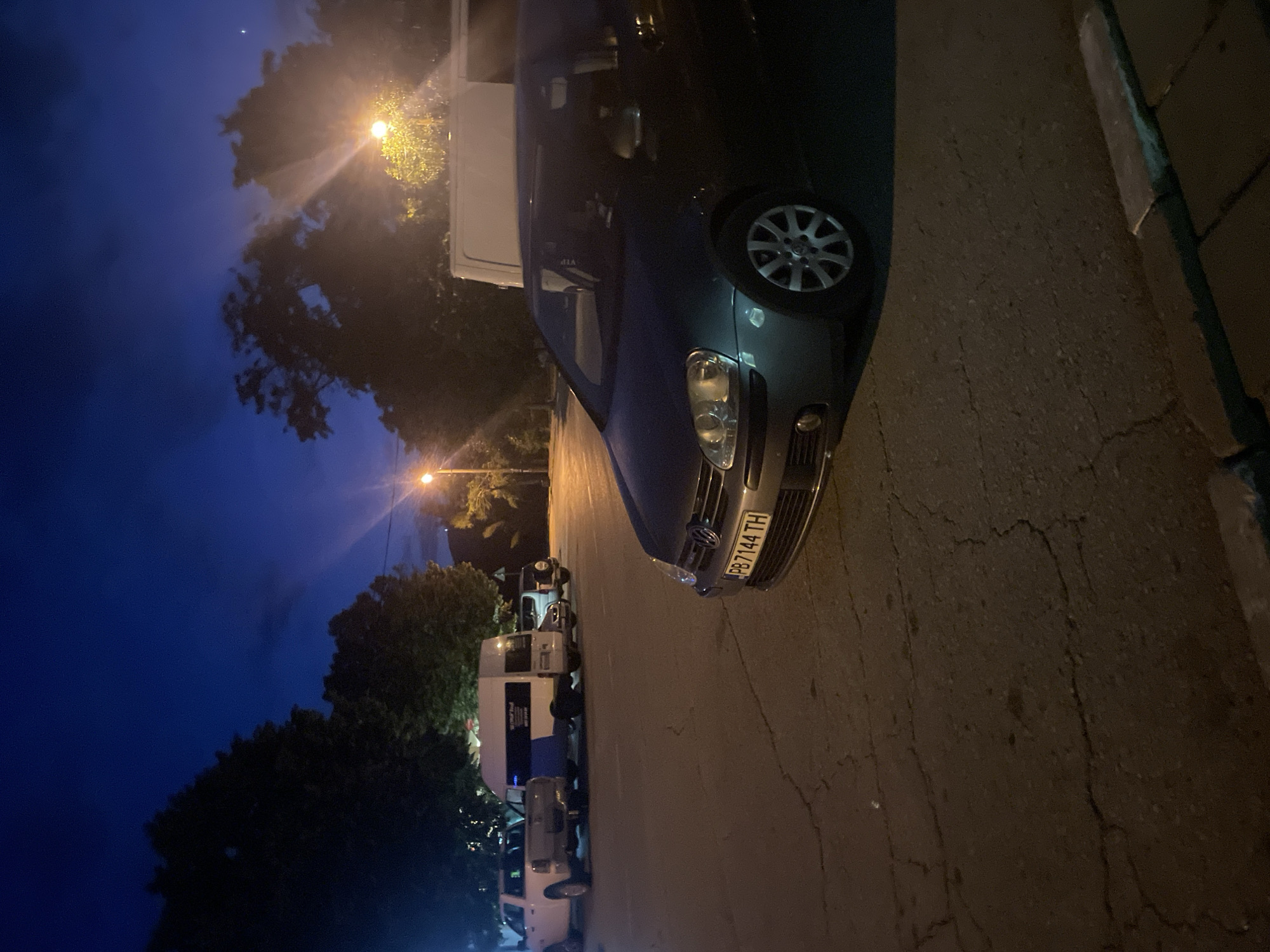 The image depicts a nighttime scene in a city, possibly Muldava, Bulgaria, characterized by a dark blue sky with some clouds illuminated by streetlights. In the foreground, the front portion of a dark-colored Volkswagen Golf is visible, with its license plate reading "PB7144 TH." The car is parked on asphalt pavement with visible cracks. Behind the Golf, a white van with blue accents is parked, and to its left, a white pickup truck is also visible. Further back, other vehicles and trees are dimly illuminated by streetlights. The overall environment suggests a parking area or street at dusk or night. There are no people clearly visible in the scene.