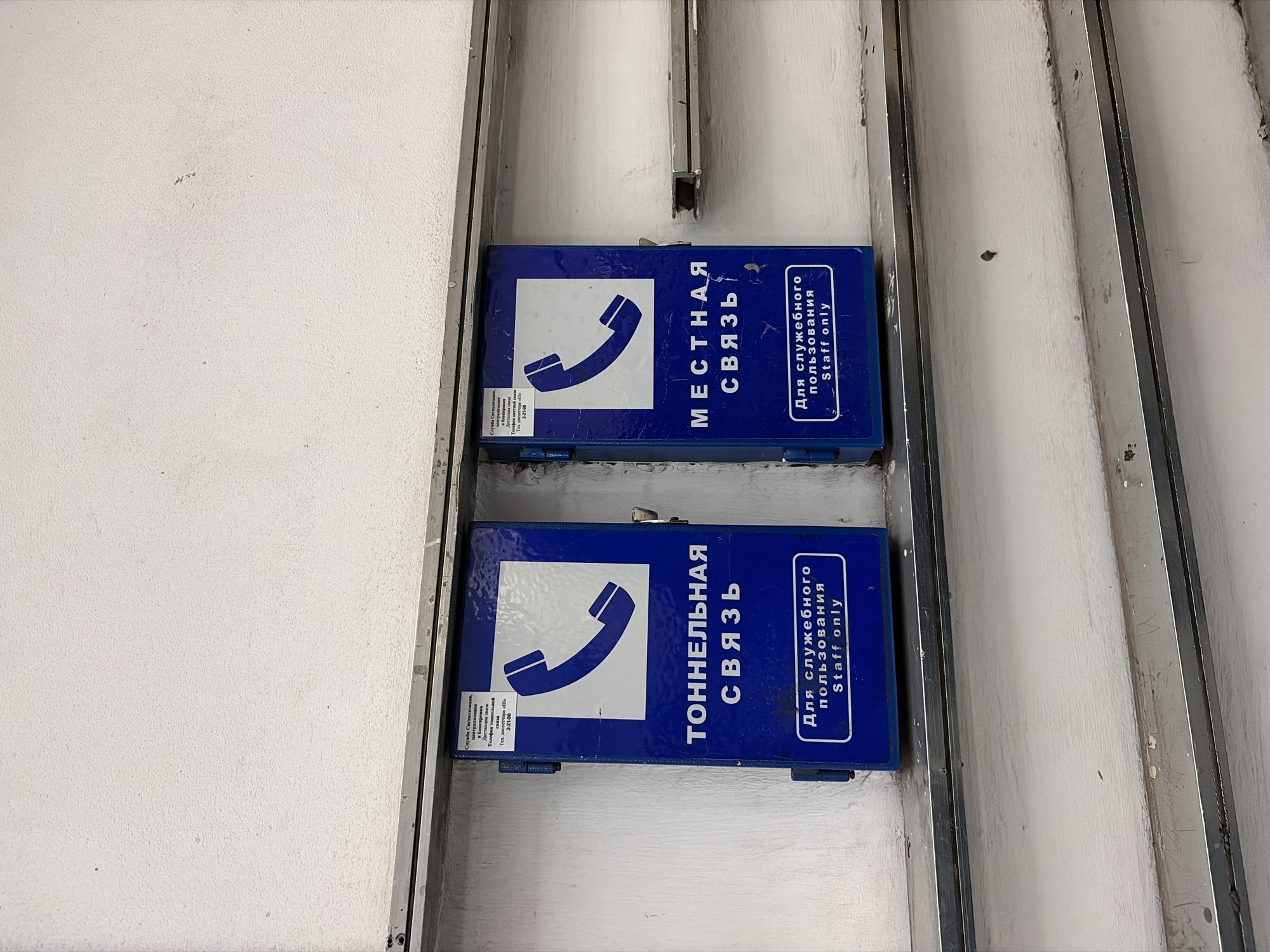 The image shows two blue boxes mounted on a white wall, likely inside a structure. Each box has a white telephone icon on the left and Russian text on the right, which translates to "Local Communication" on the top box and "Tunnel Communication" on the bottom box. Underneath the Russian text on both boxes is "For Staff Only" in English.
The scene is most likely in a tunnel or service area in Saint Petersburg, Russia, due to the presence of the Russian text and the "Tunnel Communication" box. The presence of two boxes suggests different communication systems for local and tunnel purposes. The overall environment seems industrial or utility-focused, based on the wall material and the functional design of the boxes.