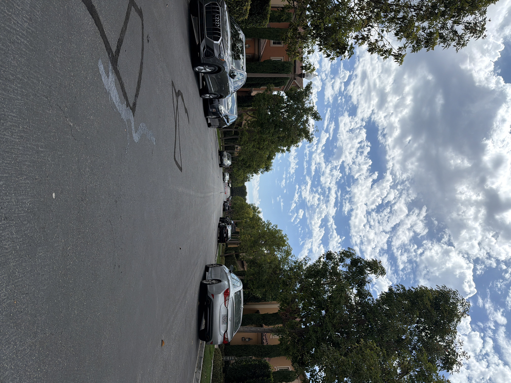 The image depicts a street scene in Irvine, California, under a partly cloudy sky. The perspective is low, close to the asphalt road which shows some dark tar repairs and what appears to be a light-colored spill. On the left side of the road, a dark grey Audi SUV is parked. Further down the street on the left, several other cars are parked, and trees line the sidewalk. The houses on the left are Mediterranean-style, with orange stucco exteriors and red-tiled roofs. On the right side of the road, a silver sedan is parked. Beyond it, more trees and houses are visible. The street is relatively narrow, and appears to be a residential area.