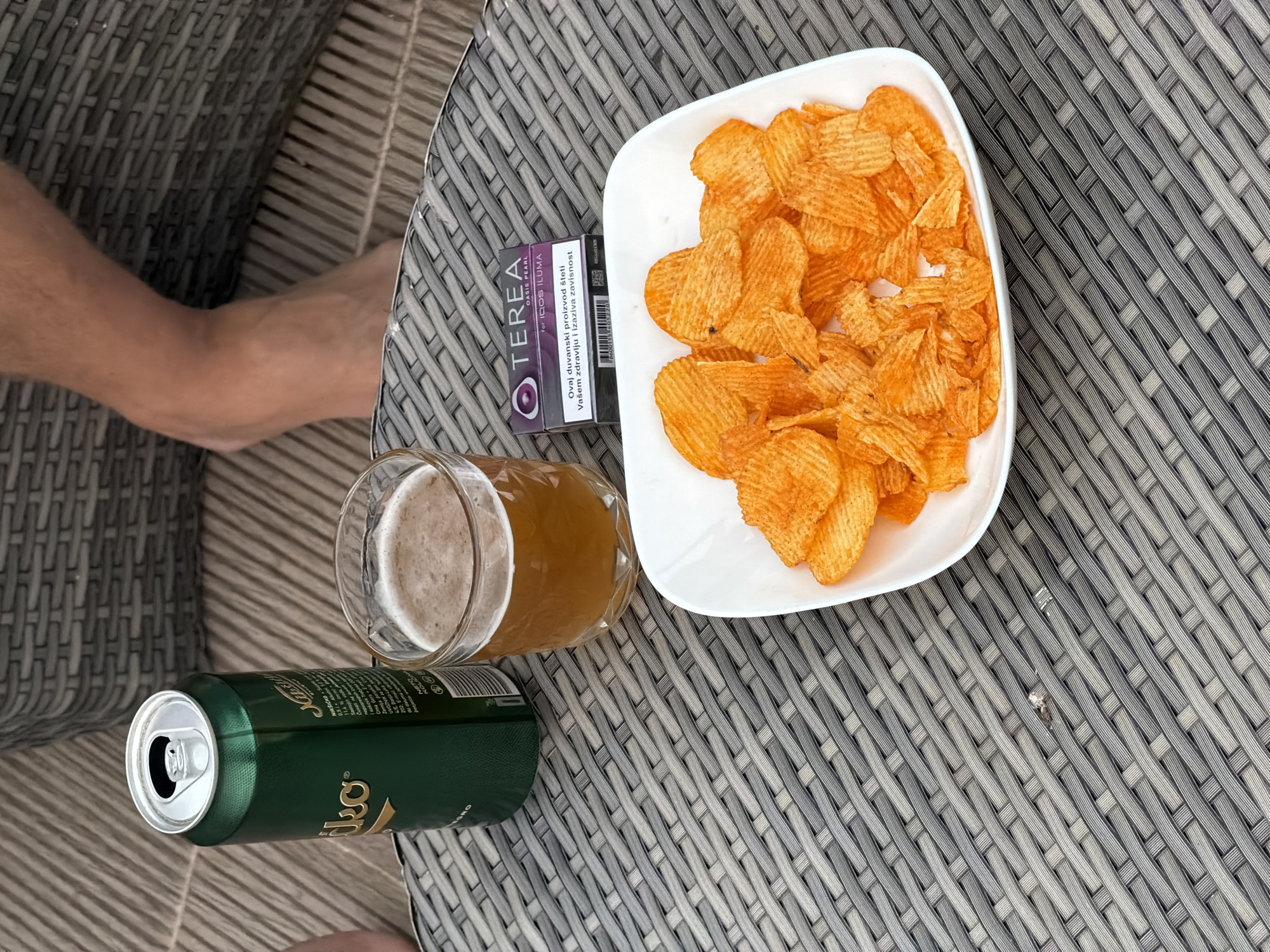 The image shows a table with a can of Nikšićko beer, a glass of beer, a pack of Terea Oasis Pearl for IQOS Iluma, and a plate of potato chips. The table has a gray woven surface. A person's leg is visible in the background. The pack of Terea Oasis Pearl has a warning stating that the product is harmful to health and causes addiction. The Nikšićko beer can has some text on it. The table appears to be outdoors. This image was taken in Budva, Montenegro.