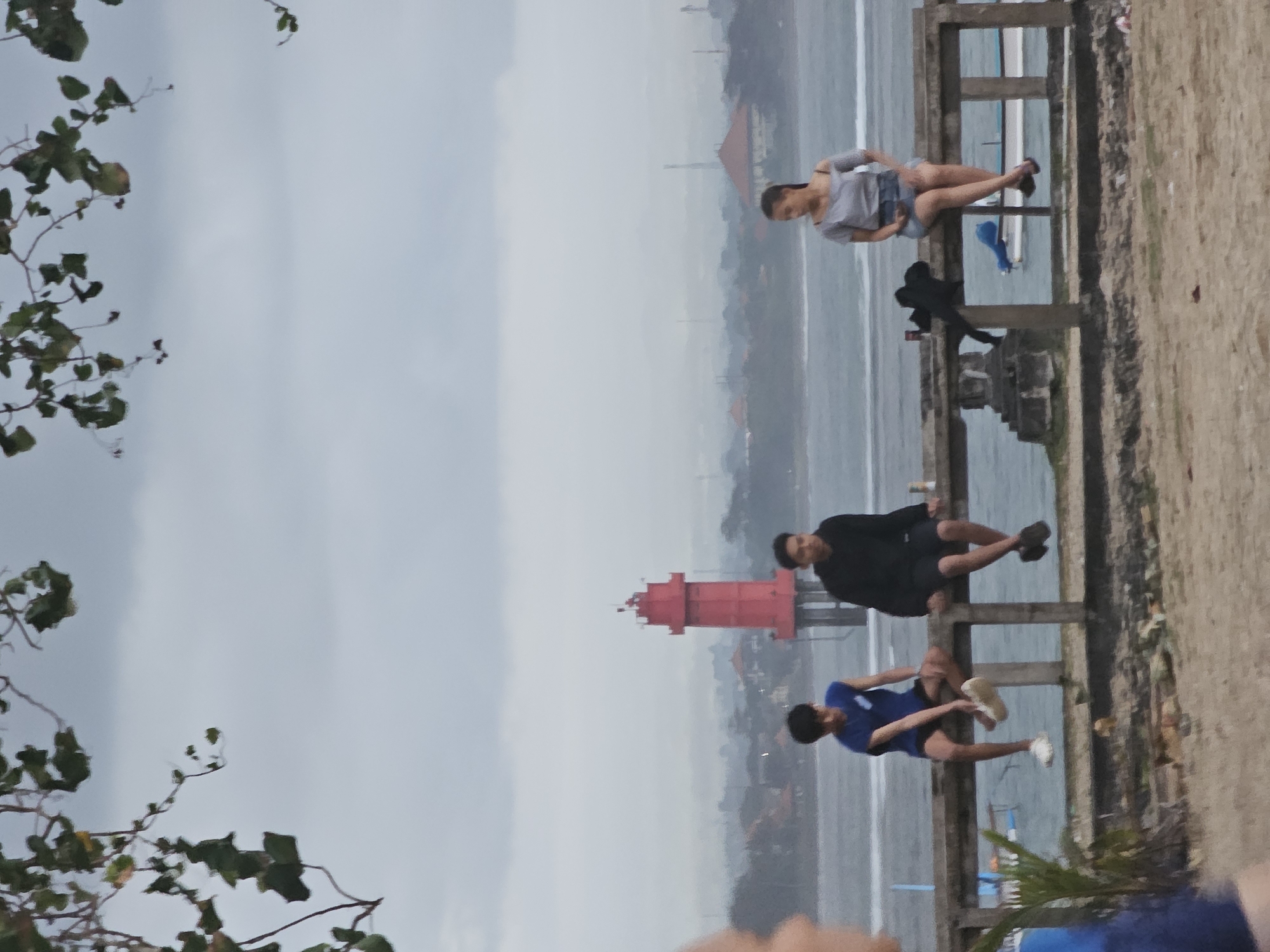 Three young adults are sitting on a concrete barrier overlooking a body of water. A red lighthouse is visible in the background, along with buildings and industrial structures, suggesting an urban coastal setting within Denpasar, Indonesia. The sky is overcast.
The person on the left is a male wearing a blue sleeveless shirt and dark shorts, with white sneakers on his feet. He is sitting with his legs dangling over the barrier.
The person in the middle is a male wearing a black hooded sweatshirt and dark shorts. He is sitting with his legs dangling over the barrier.
The person on the right is a female wearing a light gray off-the-shoulder top and denim shorts. She is sitting with her legs dangling over the barrier.
On the barrier in front of the female is a black bag. To the left of the bag is a red can, possibly a beverage can.
The foreground consists of a sandy area with some sparse vegetation. In the background, boats are visible on the water. The overall atmosphere is casual and relaxed.