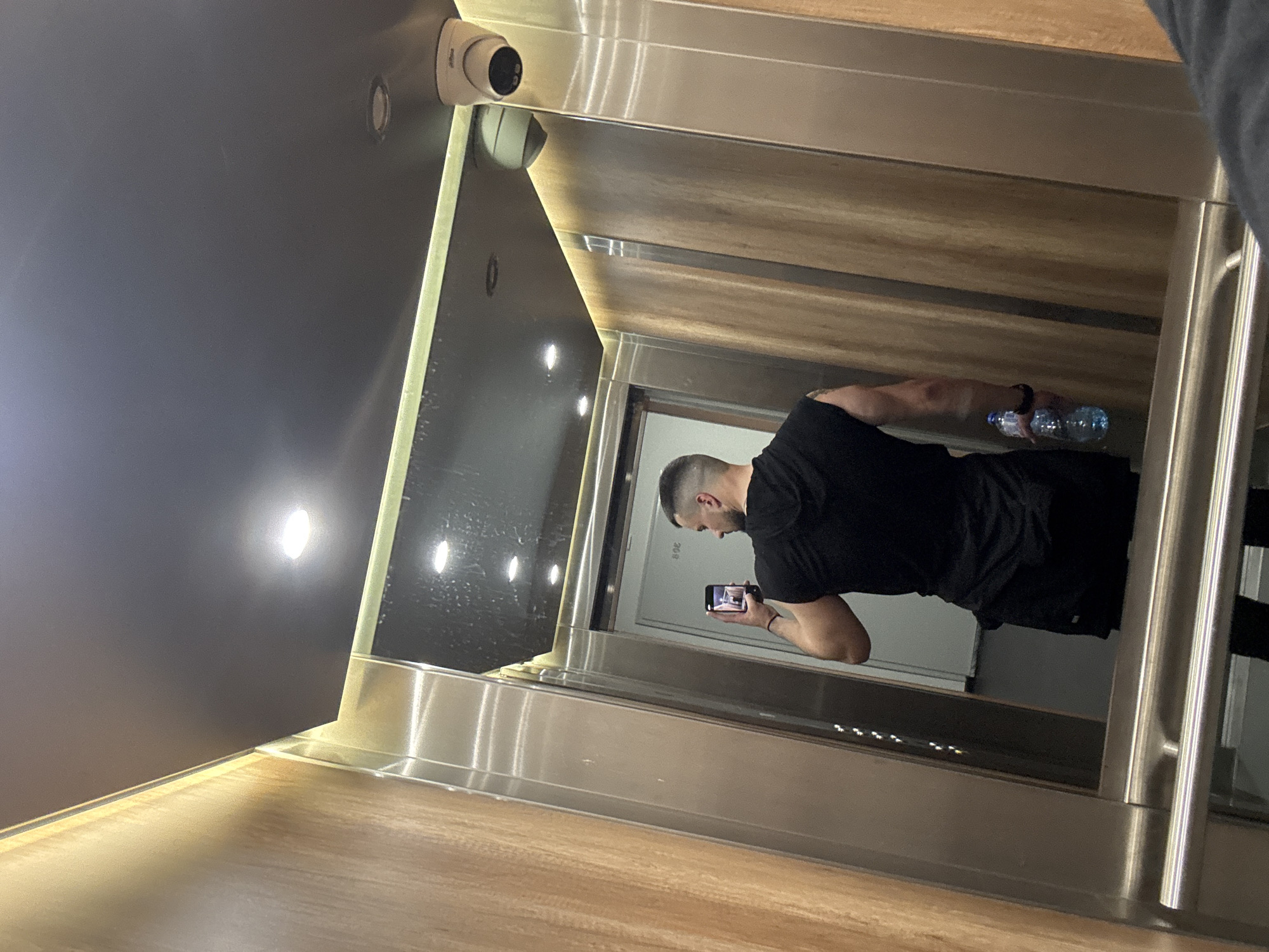 The image captures a man inside a modern elevator, facing away from the viewer, appearing to take a photo or video with his smartphone. The man, with short dark hair, is wearing a black short-sleeved shirt and black shorts. He holds a clear plastic water bottle in his left hand and a black smartphone in his right hand, pointed towards the reflective surface of the elevator.
The elevator interior features a combination of materials: a dark, possibly reflective, ceiling with several recessed circular lights, and a wall with light-colored wooden panels on the right. The left side and bottom section of the elevator are clad in polished metallic surfaces, which reflect the interior lights and the man. A metallic handrail runs along the right side of the elevator.
Mounted on the ceiling/wall junction in the upper left of the frame are two surveillance cameras, one of which clearly displays the "dahua" brand logo. The reflections on the metallic surfaces show elements of the elevator interior and the man. Faintly visible in the reflection on the door ahead of the man is the number "308". The lighting is bright and even, originating from the recessed ceiling lights. The overall setting suggests a contemporary building in Sofia, Bulgaria.