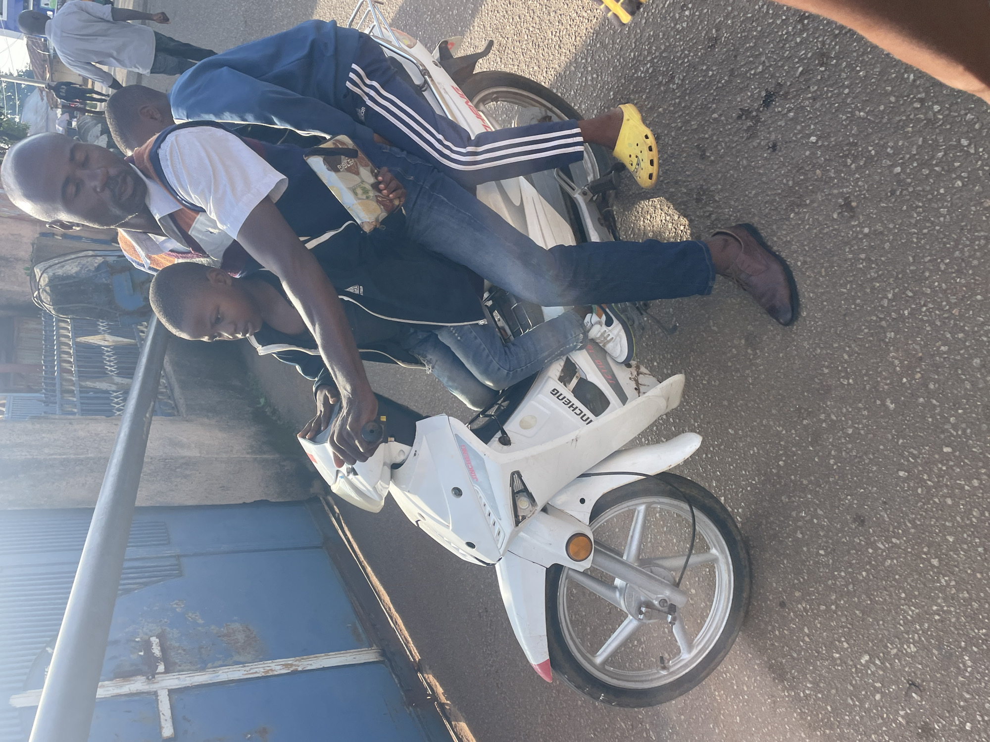 A man wearing a blue and white striped vest over a white shirt and blue jeans is riding a white motorbike. He is carrying two children. A boy in a dark jacket is seated in front of him, and another boy in a blue sweatshirt is seated behind him. The motorbike is traveling on a paved road with a slight incline. To the left of the motorbike is a blue gate with a metal railing, and to the right is a textured concrete wall. In the background, other people and vehicles are visible. The scene appears to be in an urban environment, possibly Jos, Nigeria, given the context. The lighting suggests it is daytime.