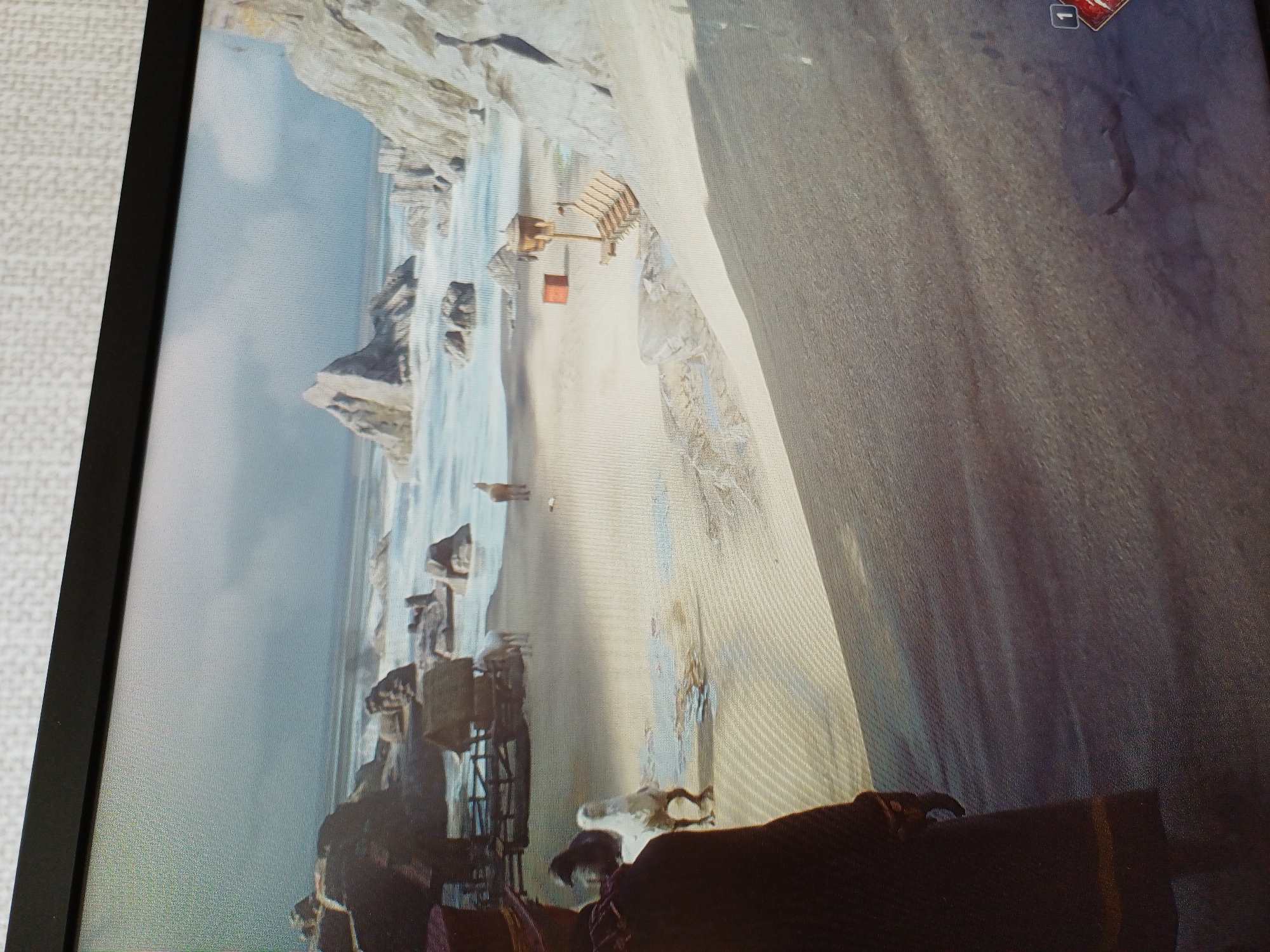In the city of Unknown, Unknown, a scene unfolds on a sandy beach with rocky outcrops and the ocean in the background. The environment is overcast with a light gray sky. In the foreground, a person is viewed from behind, wearing a dark coat and a hat. To their left, a white bird stands on the sand, and a dark bird is partially visible behind it. Further into the scene, a simple wooden structure with a thatched roof sits on the beach. Two horses are near this structure, and another animal, possibly a deer or llama, stands further out on the sand. To the right, a small wooden structure with a birdhouse-like top is mounted on a post. Beside it, a wooden object resembling a lounge chair is placed on the sand. A red cylindrical object is also present near the wooden structure. The sand is textured, with some areas appearing wet or rippled. A small red indicator with the number "1" is visible in the lower right corner of the image.