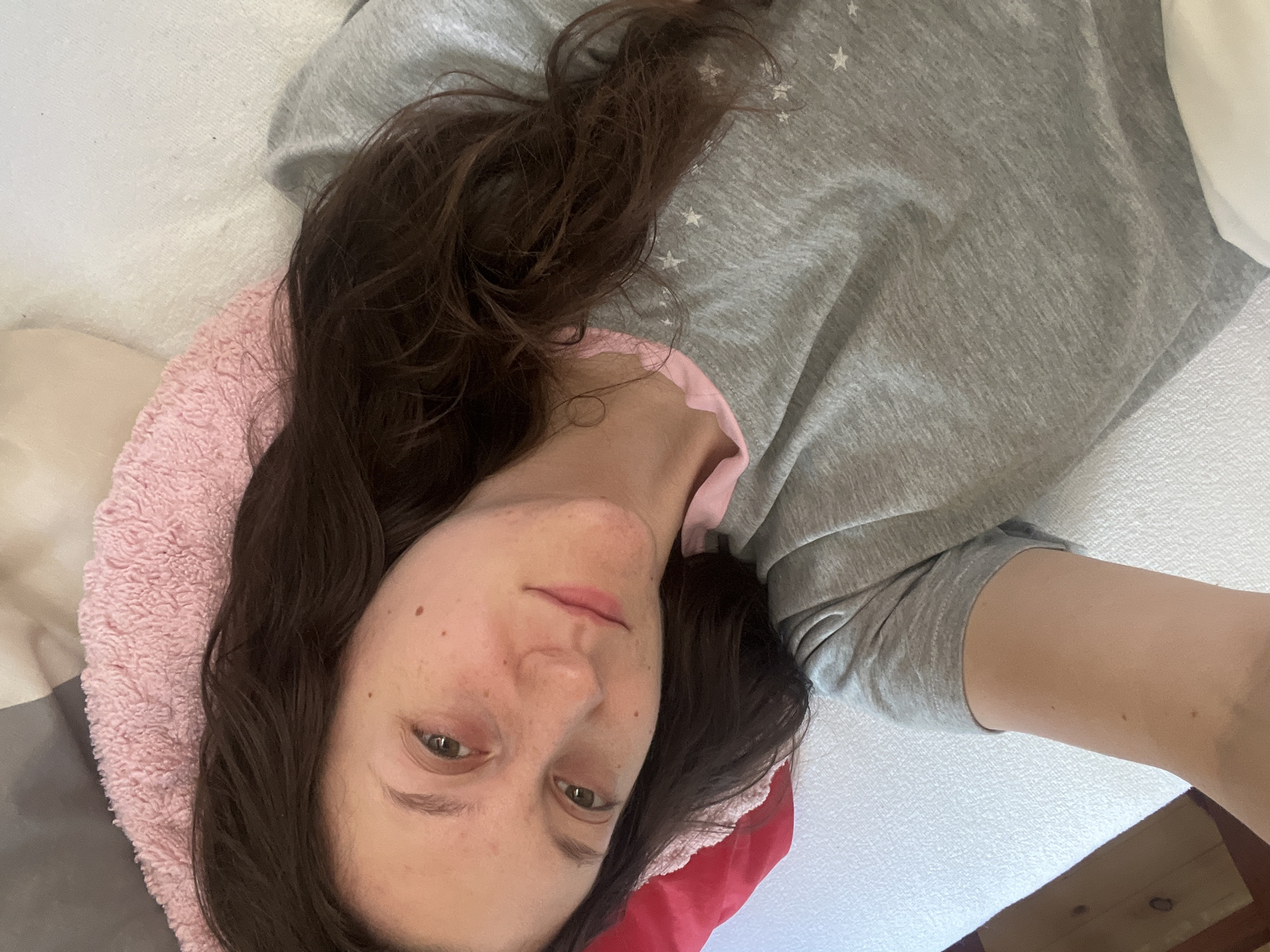 In Popintsi, Bulgaria, a young woman with long, dark brown hair is lying down, looking at the camera. She is wearing a grey t-shirt with small white stars on it and a pink trim around the collar. Her left arm is resting on a white surface, and her head is propped up on a pink fuzzy pillow, which is on top of a grey and white patterned surface. A red pillow is visible behind her head on the left. The environment appears to be indoors, possibly a bedroom, with a white bed or couch as the primary surface. The lighting is soft, suggesting it could be daytime.