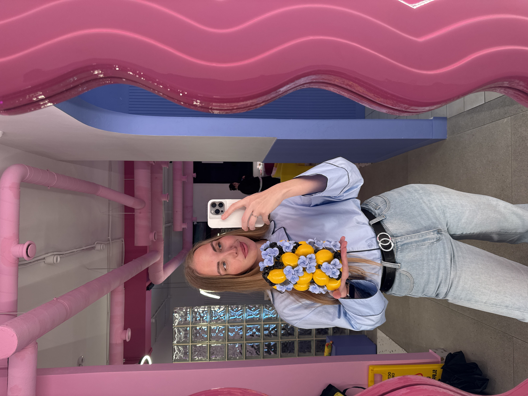 A young woman is taking a selfie in a room with playful, colorful decor. She holds up a tray of yellow, lemon-shaped treats decorated with blue flowers and blackberries. The background features pink and blue accents, including decorative pipes and a wave-shaped ceiling detail, all contributing to a whimsical atmosphere. The woman is wearing a light blue shirt and jeans, accessorized with a black belt. The scene is a reflection in a mirror, suggesting the woman is inside a store or cafe. According to the metadata, the location is Sochi, Russia.