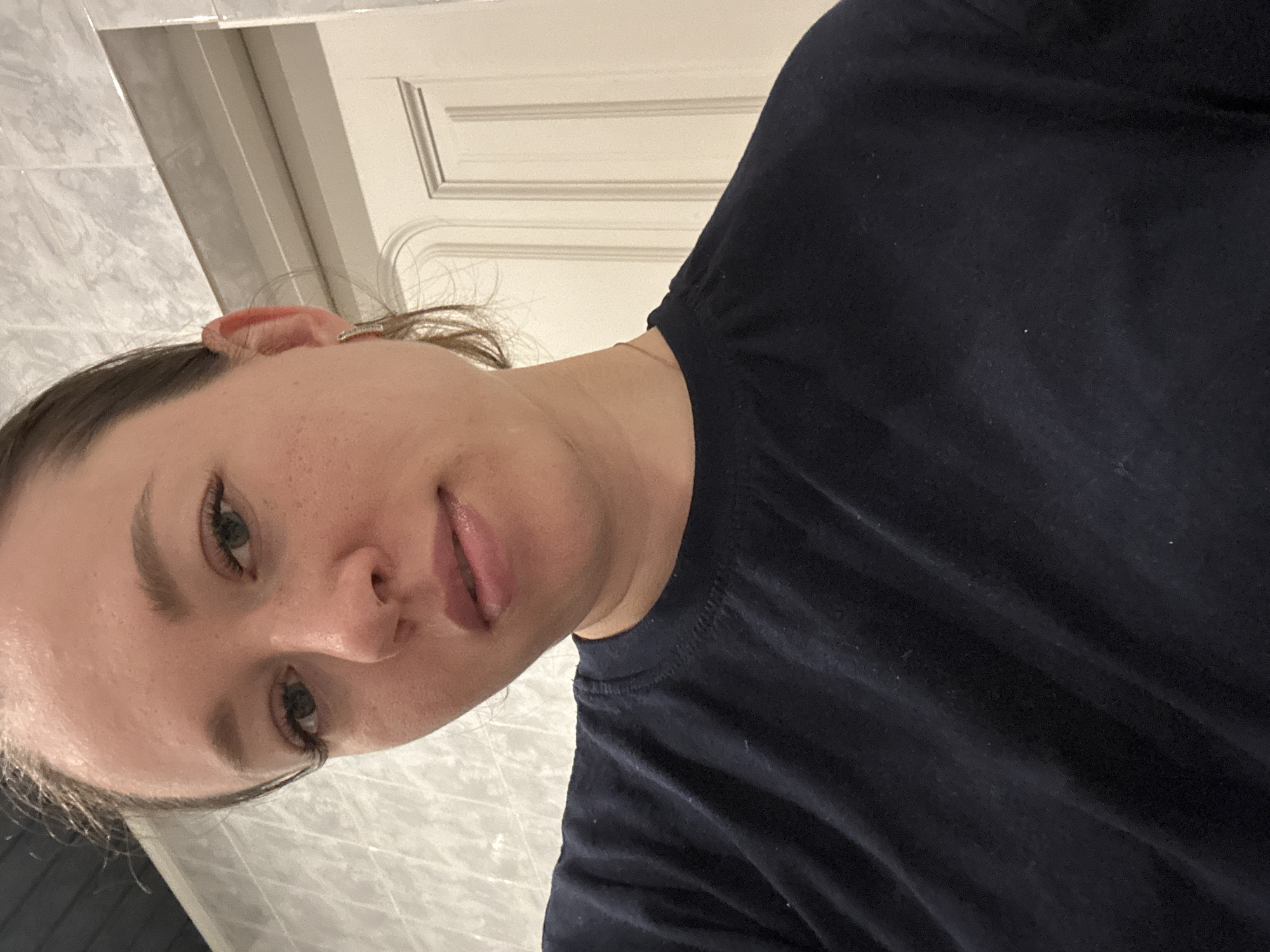 A woman wearing a dark blue or black shirt is facing the camera. The background shows what appears to be a bathroom or tiled area. A partially visible door is also in the background. The image is rotated. The location is not clearly indicated.